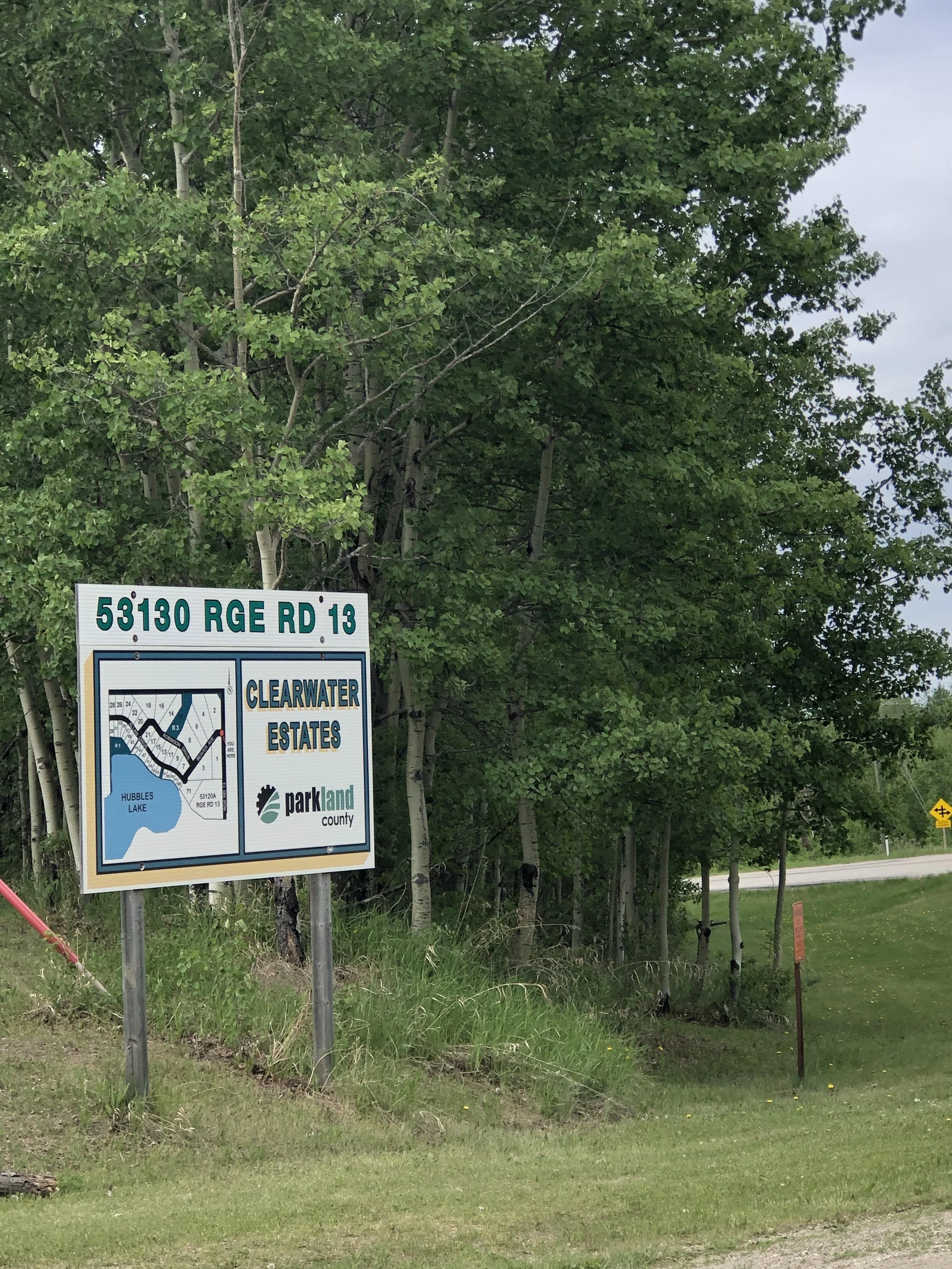A sign at a residential area reads '53130 RGE RD 13'. Behind the sign is a sign with a map of Clearwater Estates, part of Parkland County, with a background of green trees and grass, and a partly cloudy sky.