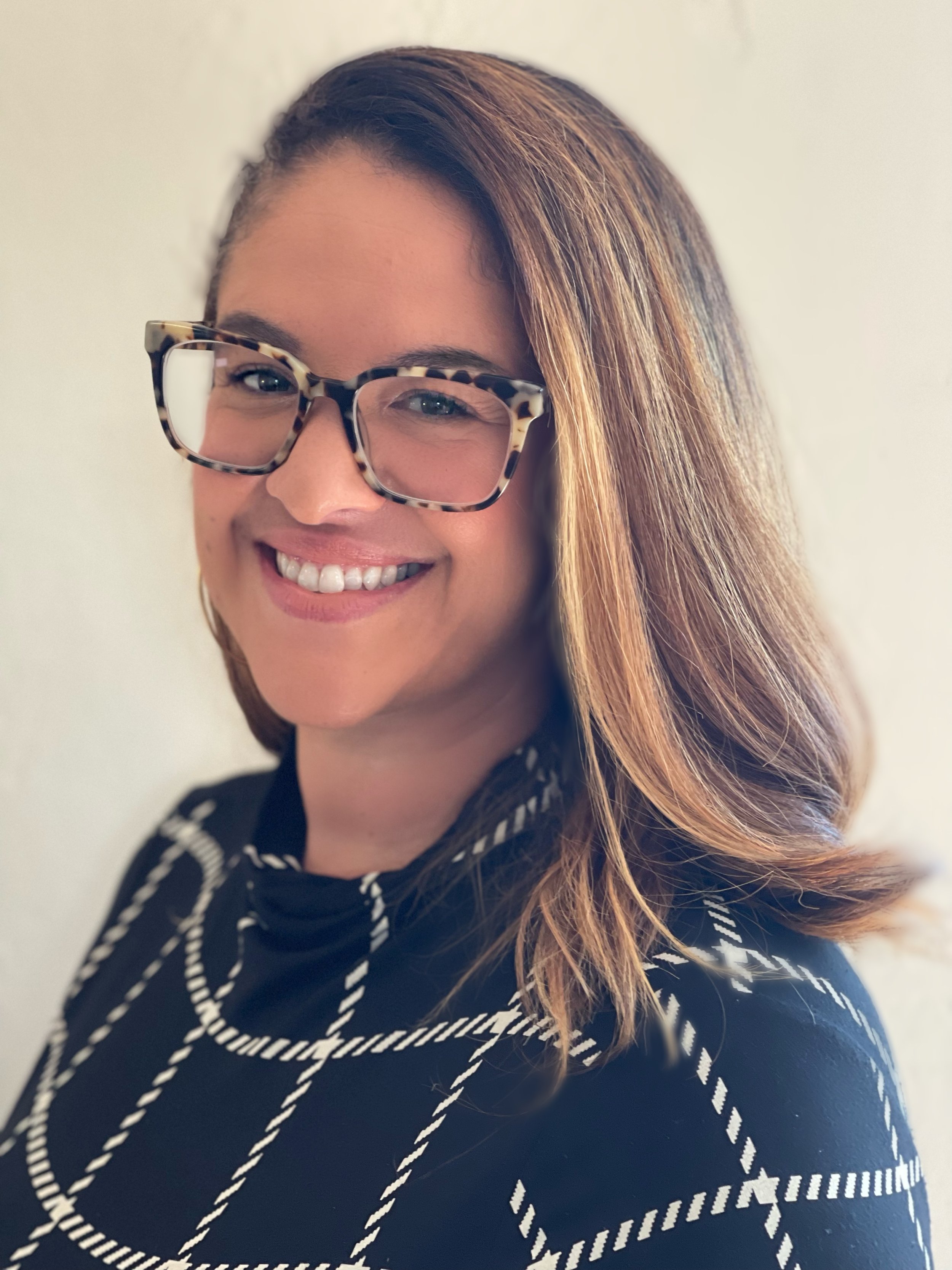 A woman with shoulder-length light brown hair, wearing large tortoiseshell glasses, smiling, dressed in a black and white striped shirt, standing against a plain light-colored background.