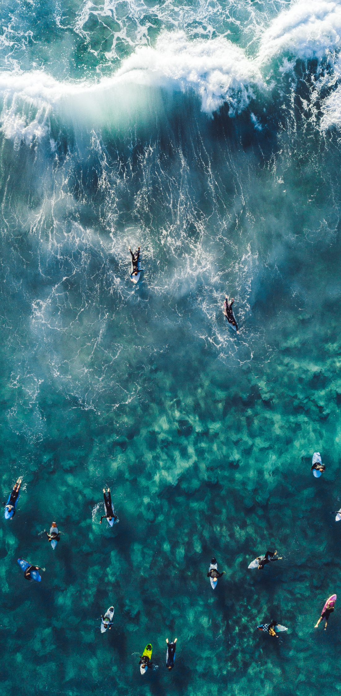 Aerial view of multiple surfers in the ocean, with a large wave crashing in the background.