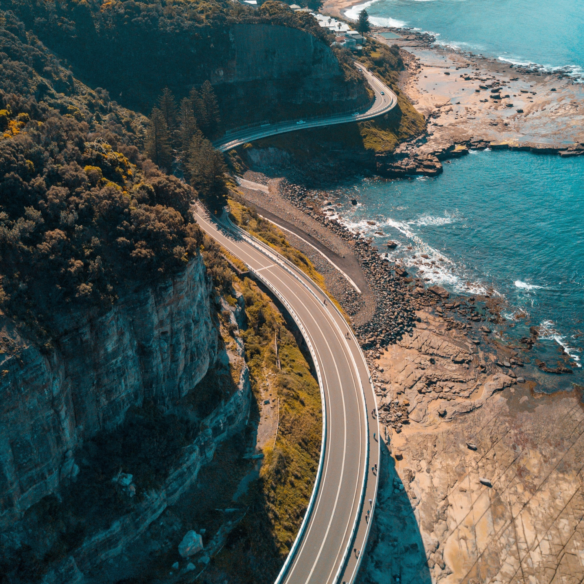 An aerial view of a coastal highway winding along the edge of a cliff, overlooking the ocean with rocky shoreline and forested hills.
