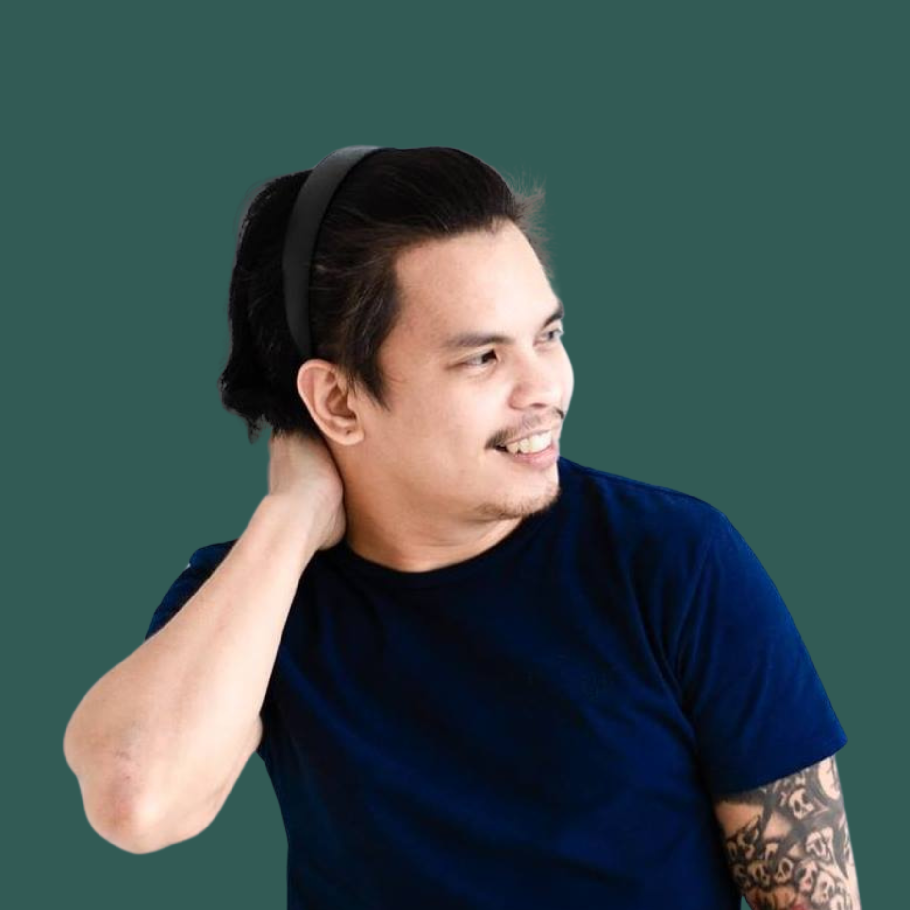 Close-up of a smiling young man with black hair, wearing a black headband and a navy blue shirt, resting his hand on his neck, with a tattooed arm visible, against a solid dark green background.