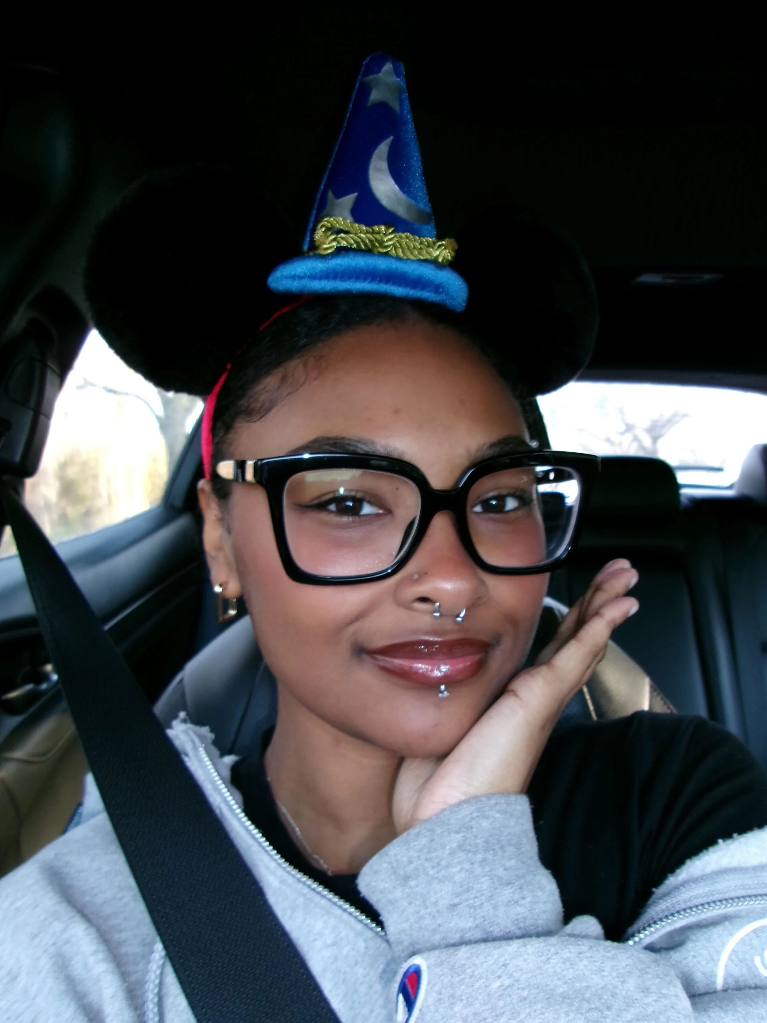 A person with glasses, multiple facial piercings, and earrings is seated in a vehicle, wearing a gray hoodie, and a Minnie Mouse ears headband with a wizard hat on top.