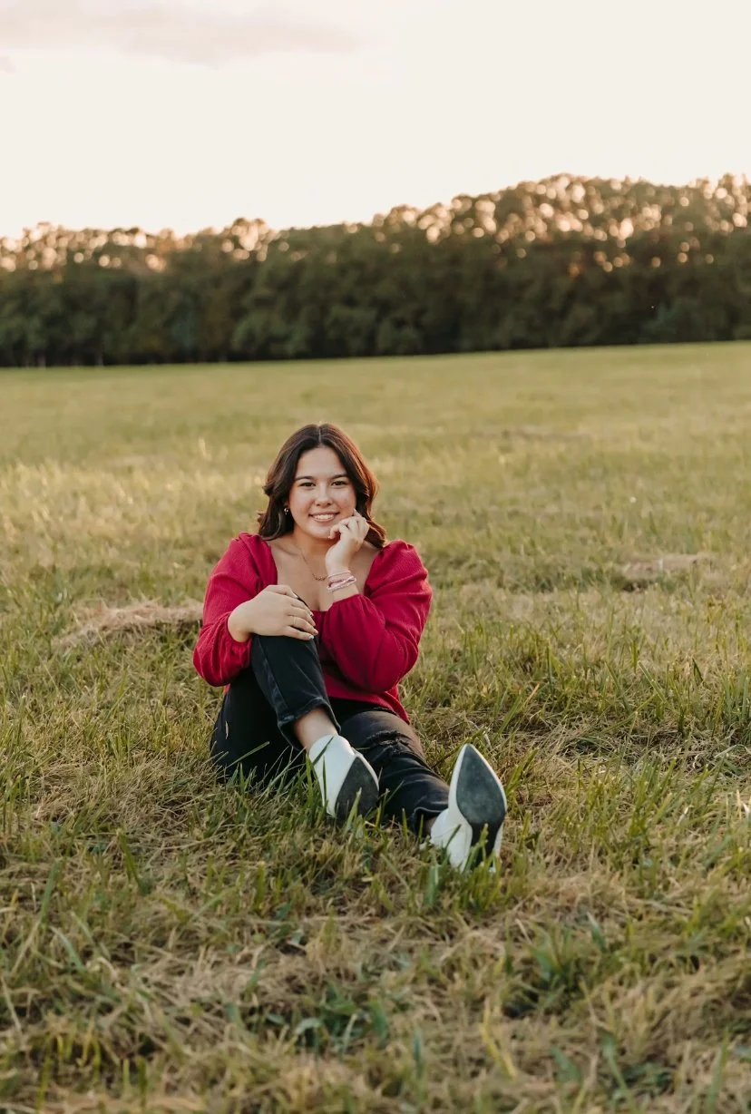 A young woman in a red sweater and black jeans sitting on grass in an open field, smiling, with trees in the background during sunset.