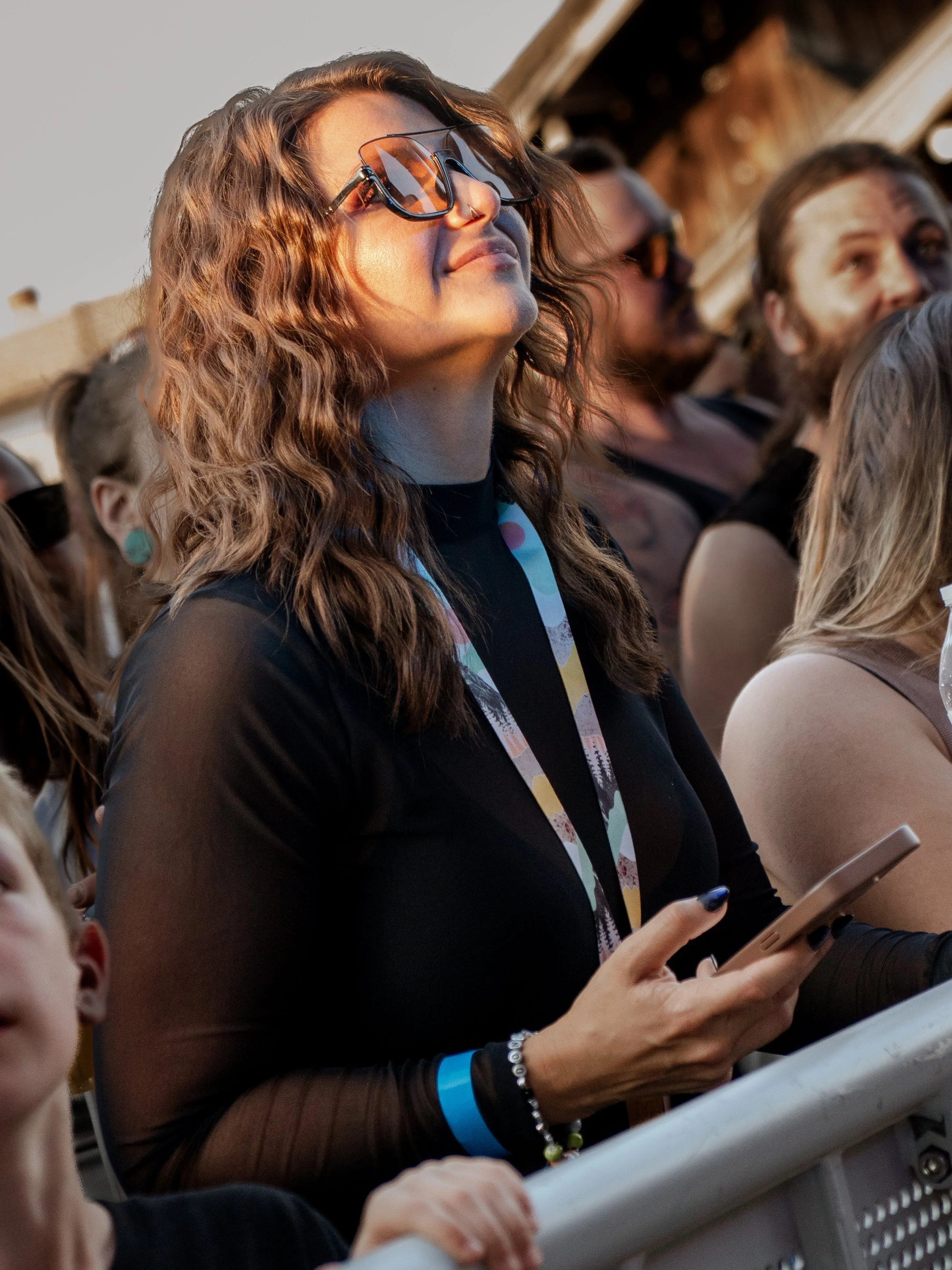 A woman with wavy brown hair and sunglasses, smiling while looking up, standing among a group of people at an outdoor event during sunset.