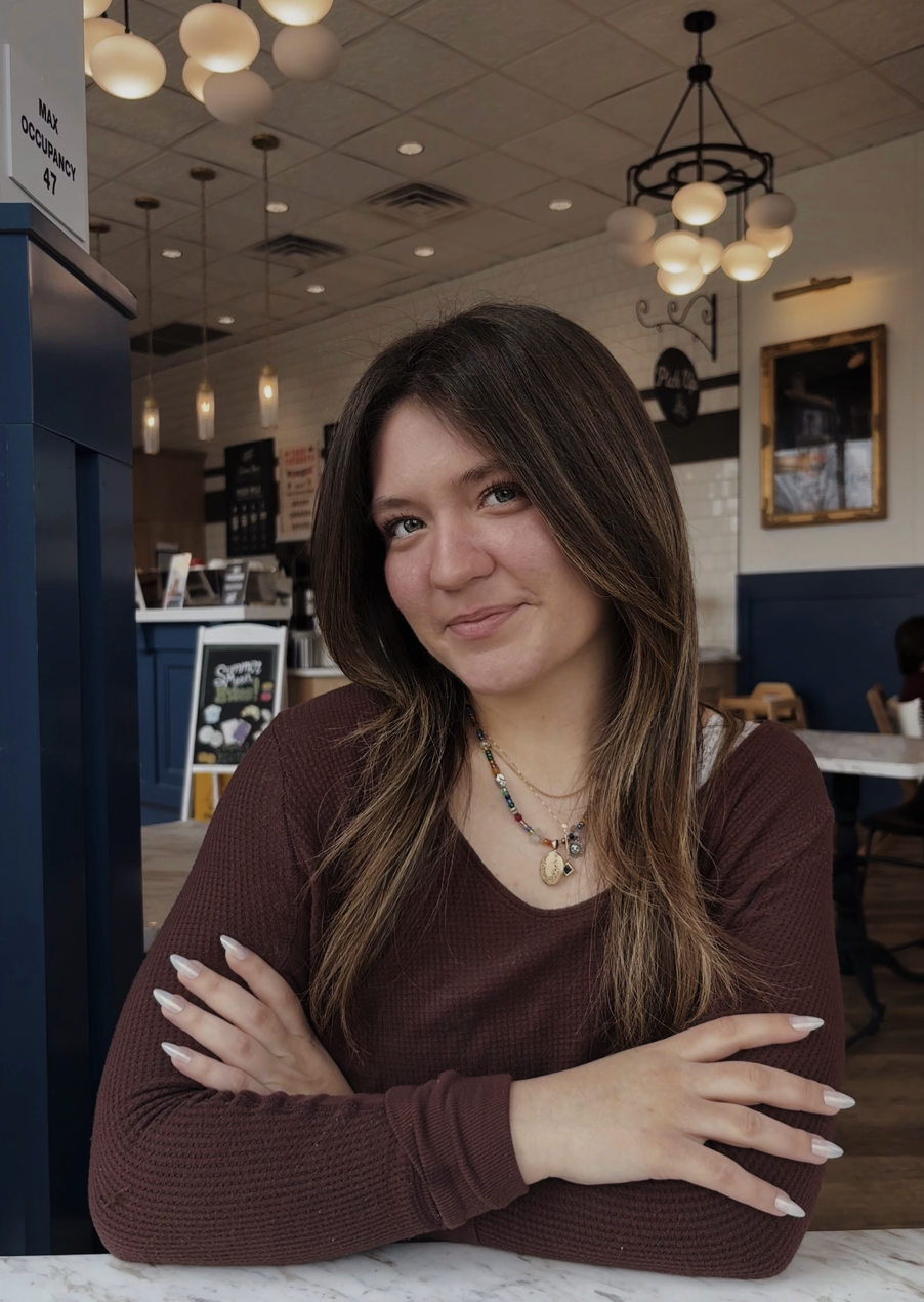 A young woman with long brown hair and light skin sits at a marble table in a cozy cafe, smiling at the camera, wearing a maroon long sleeve shirt and layered necklaces.