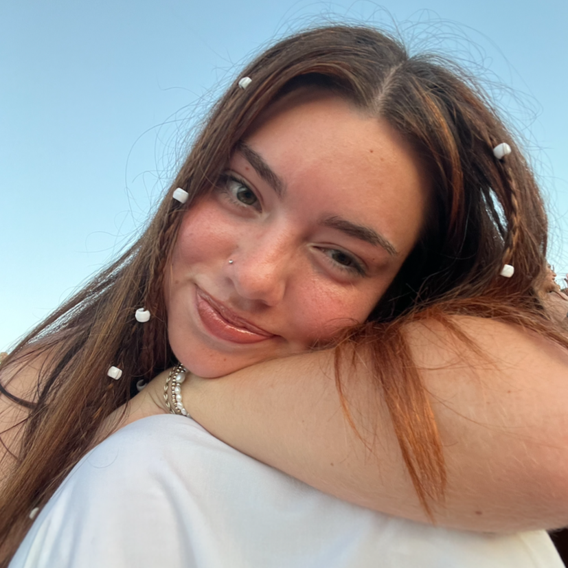 Close-up of a young woman with long brown hair and a nose piercing, smiling softly, resting her head on another person's shoulder, against a clear blue sky.
