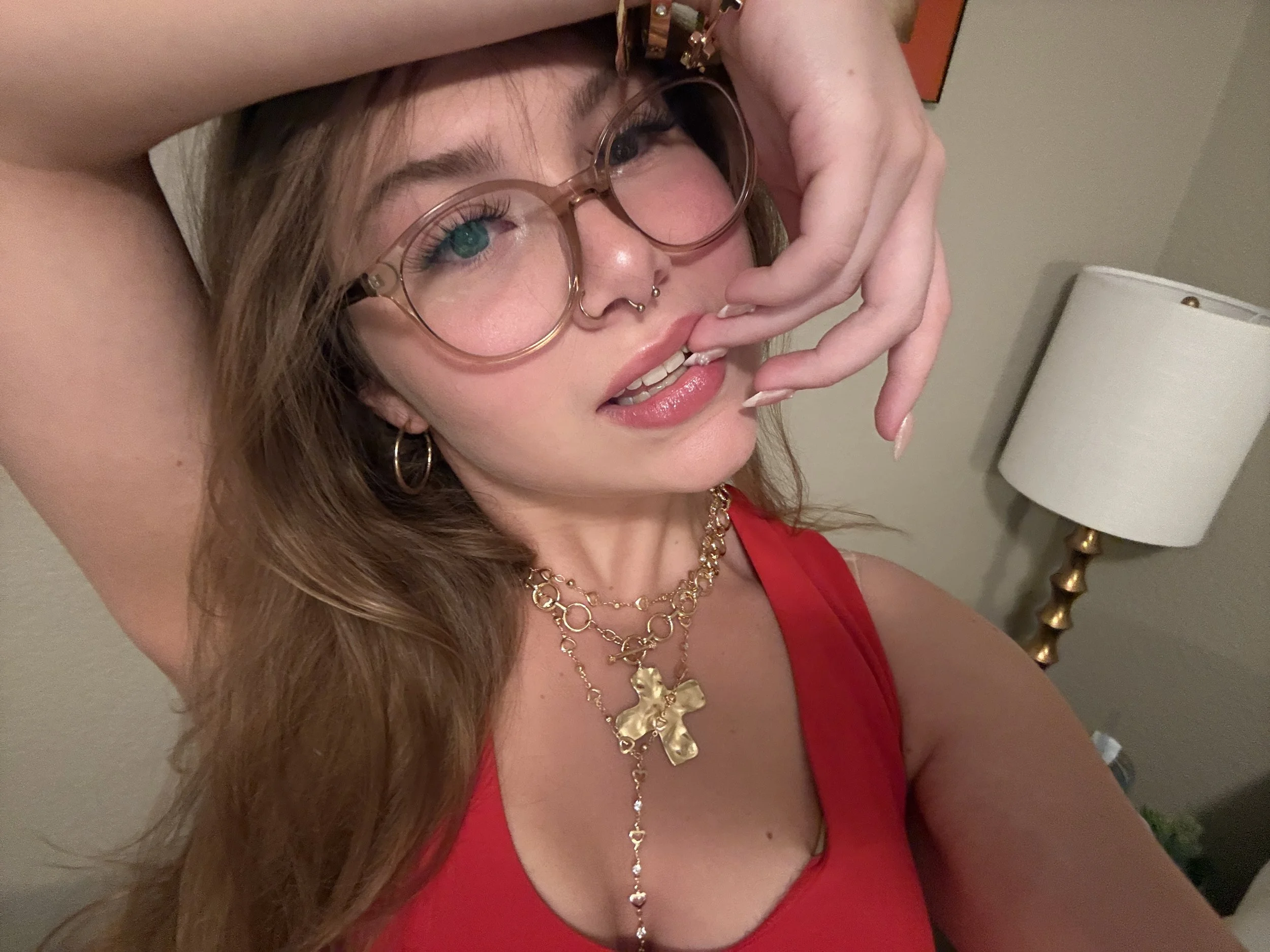 A woman with long brown hair wearing glasses, gold jewelry, and a red top, posing indoors with her hand near her face.