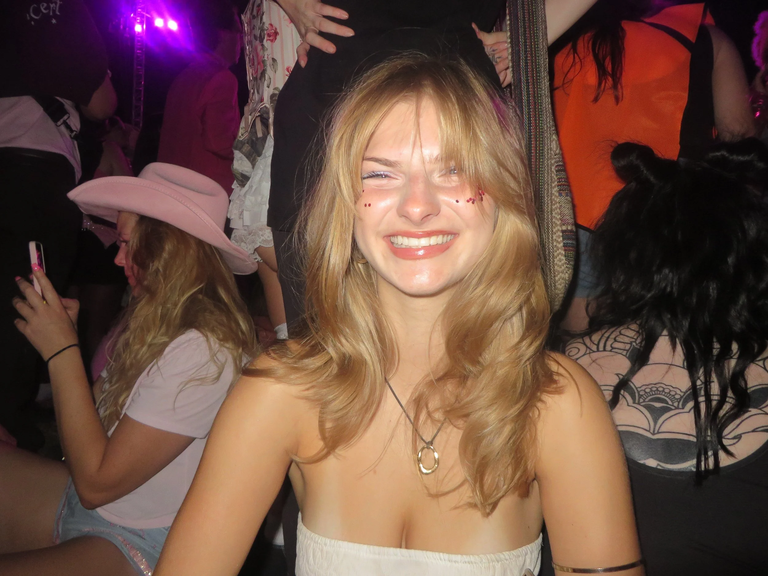 A smiling woman with blonde hair and face paint, surrounded by friends in a lively party setting.