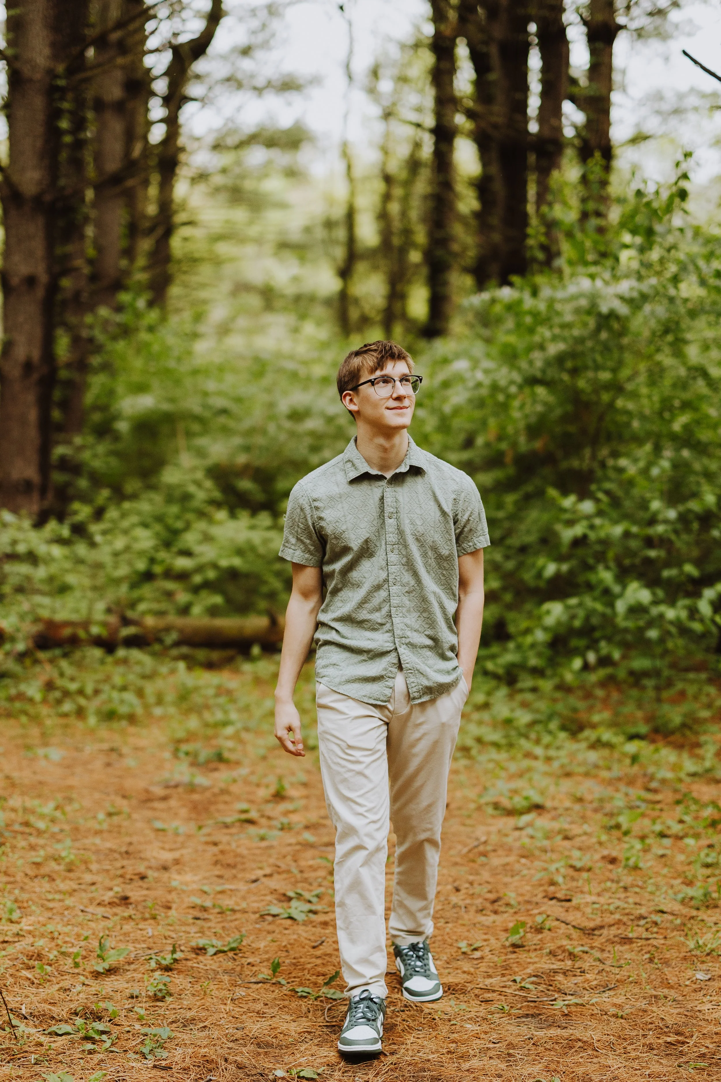 A young man with glasses, light brown hair, wearing a gray short-sleeve shirt and beige pants, walking on a forest trail with green trees and foliage in the background.