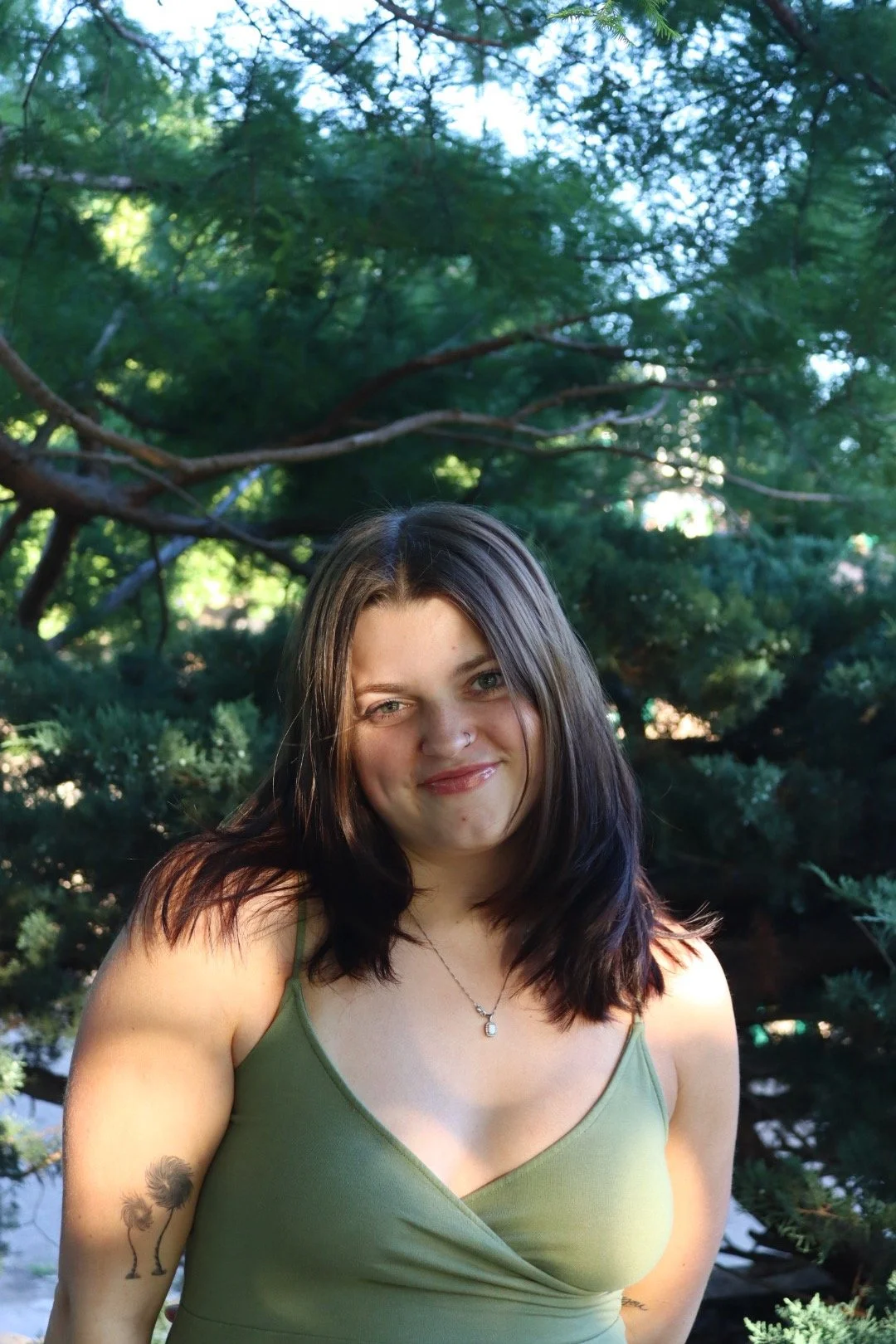 A woman with dark brown hair and a small nose piercing smiling outdoors, wearing a green tank top and a necklace, with trees and greenery in the background.