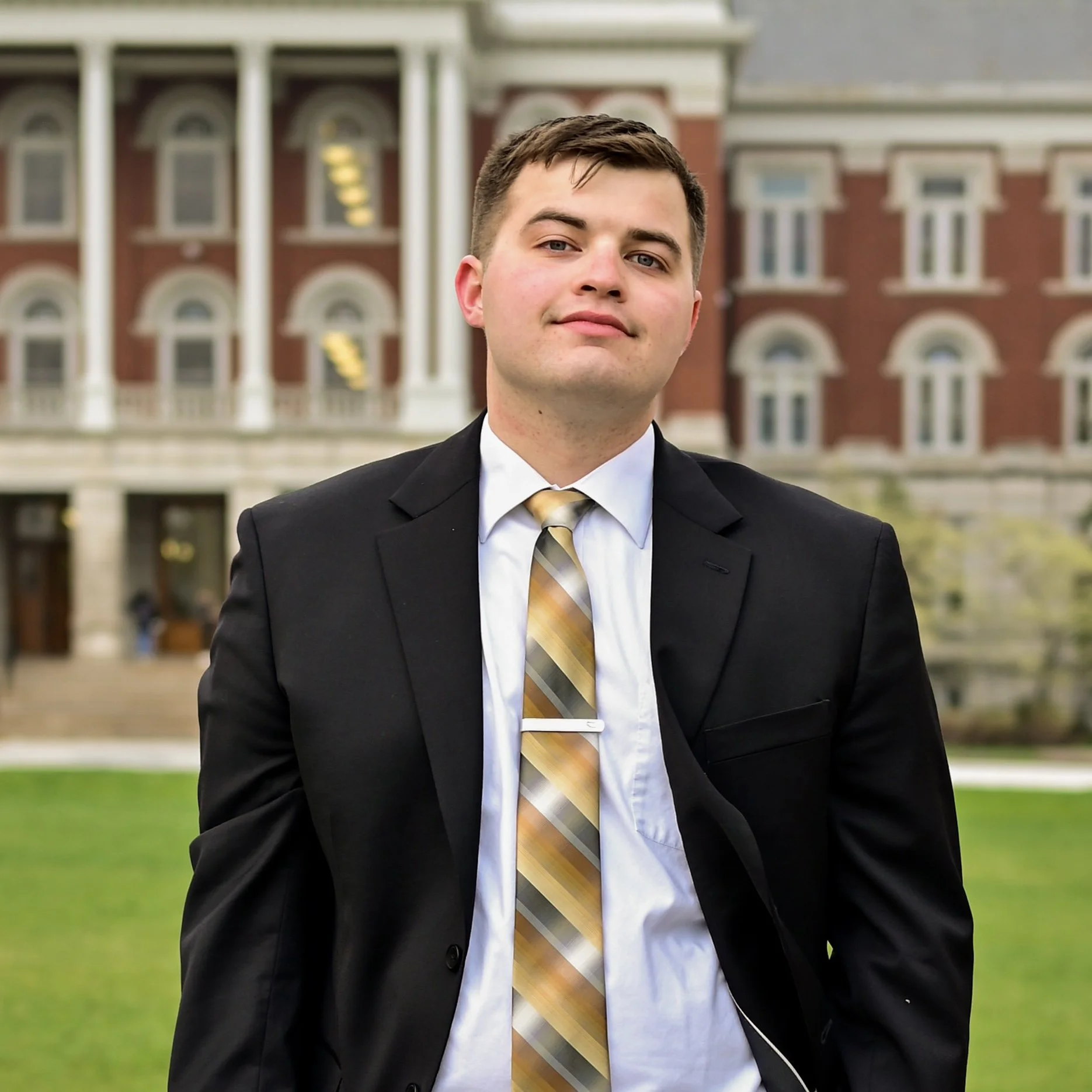 A young man wearing a black suit, white shirt, and a yellow plaid tie standing outdoors in front of a large, red brick building with white columns and arched windows.
