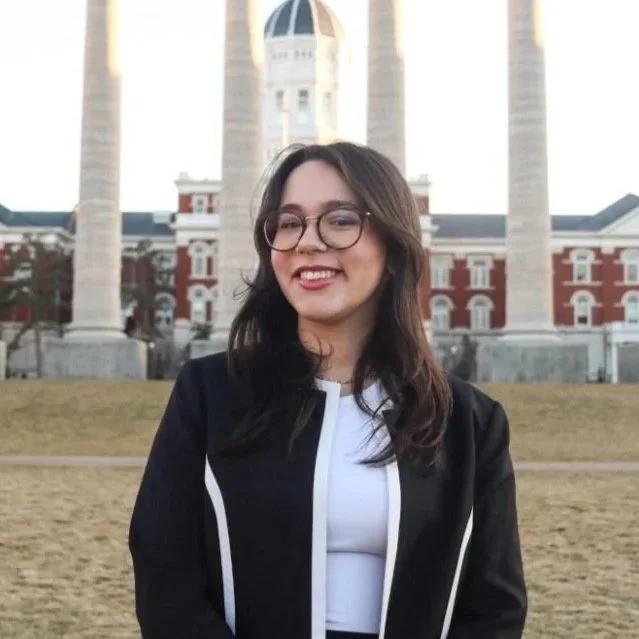 Young woman smiling, wearing glasses and a black jacket, standing in front of a historic building with columns and a dome, on a grassy area.