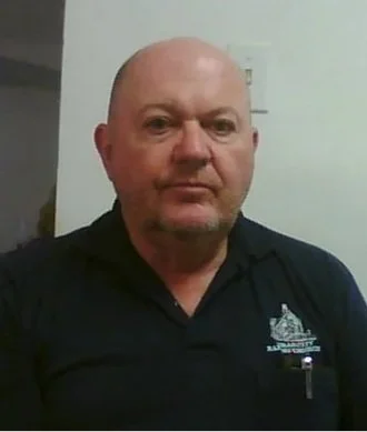 A bald man wearing a black collared shirt with an embroidered logo, standing indoors near a plain wall.