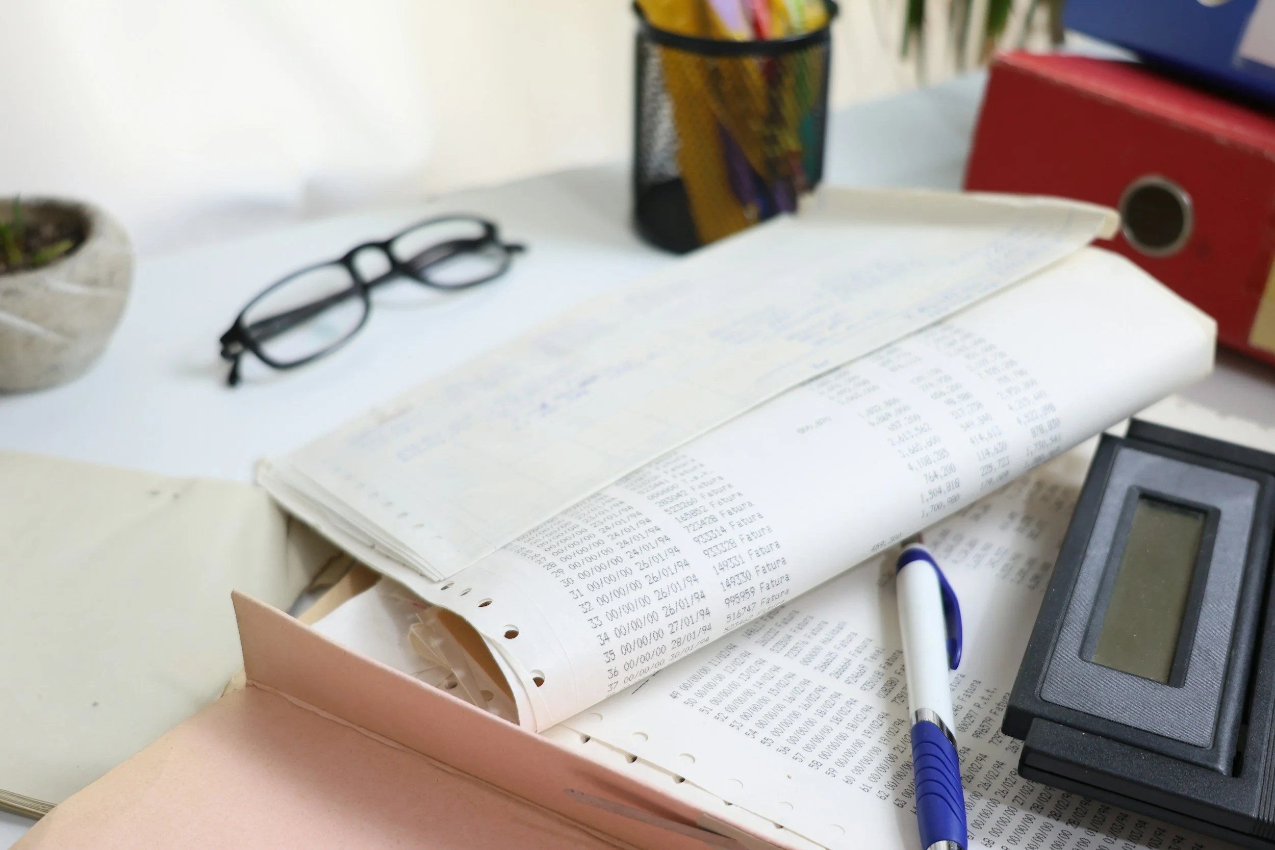 A cluttered desk surface with printed receipts, a blue and white marker, a calculator, a white surface, a pair of black eyeglasses, a colorful container holding pens or markers, and a red storage box.