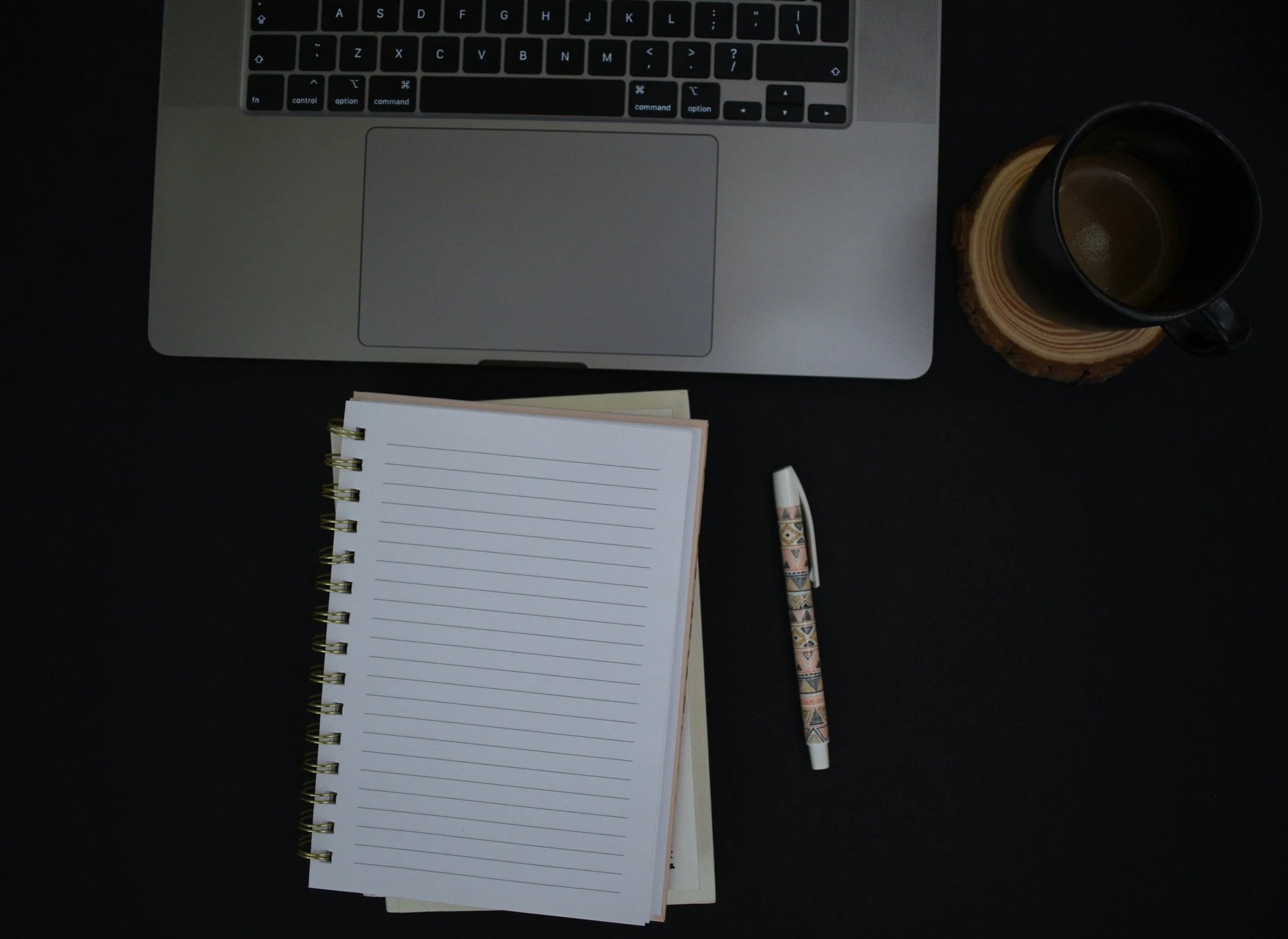 A laptop keyboard and trackpad, a spiral notebook with lined pages, a decorative pen, a black coffee mug on a coaster, and a closed book or notebook.