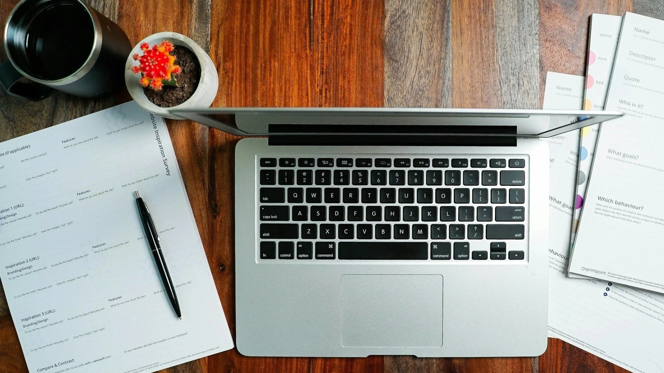 Top-down view of a workspace with a laptop, papers, a pen, a black mug, and a small potted plant on a wooden desk.