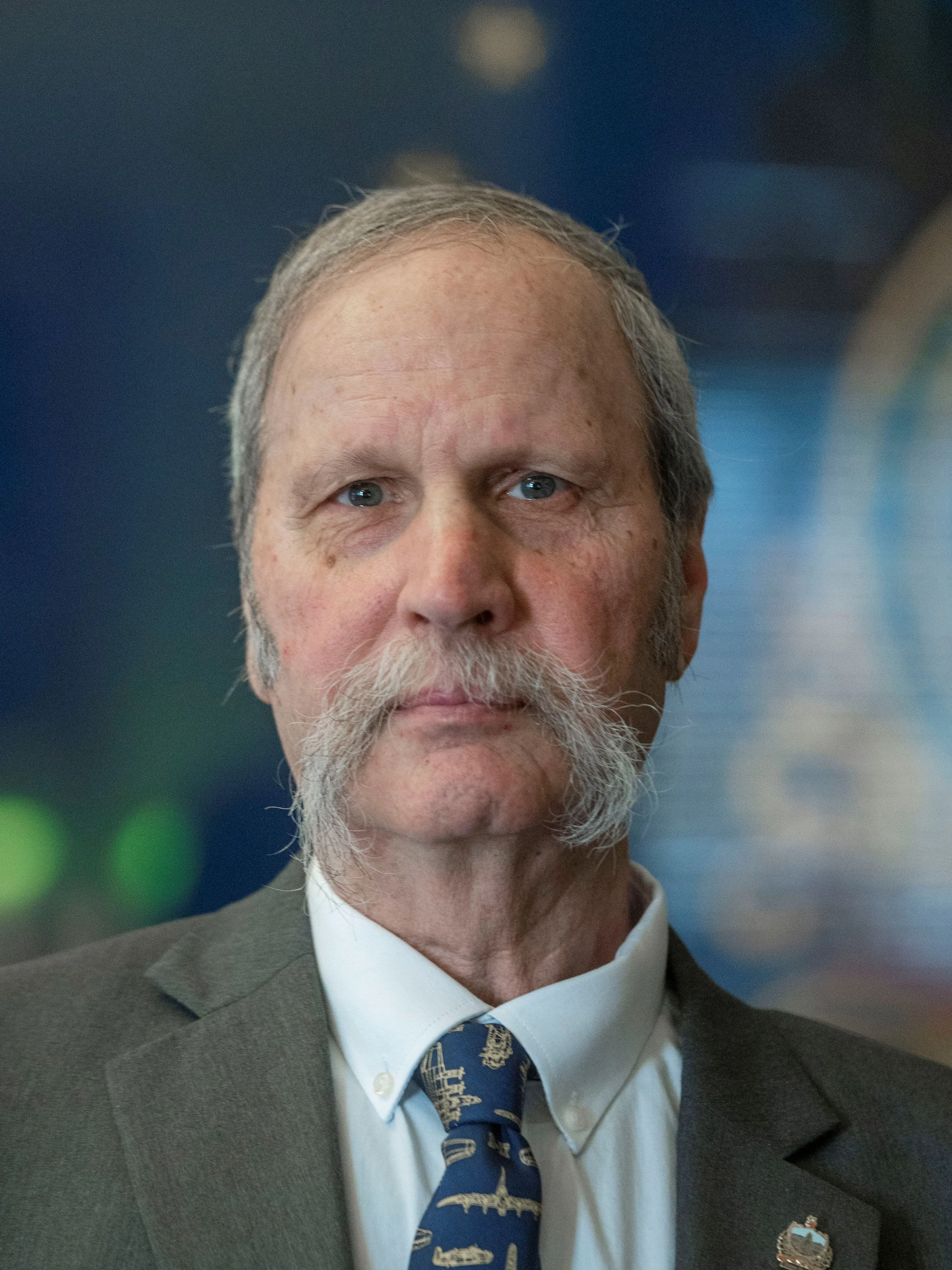A close-up photo of an older man with gray hair, a beard, and blue eyes, dressed in a gray suit, light blue shirt, and a navy tie with musical instrument patterns. He also has a lapel pin on his suit.