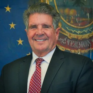 Smiling man in a suit and tie standing in front of a blue backdrop with stars and a circular emblem.