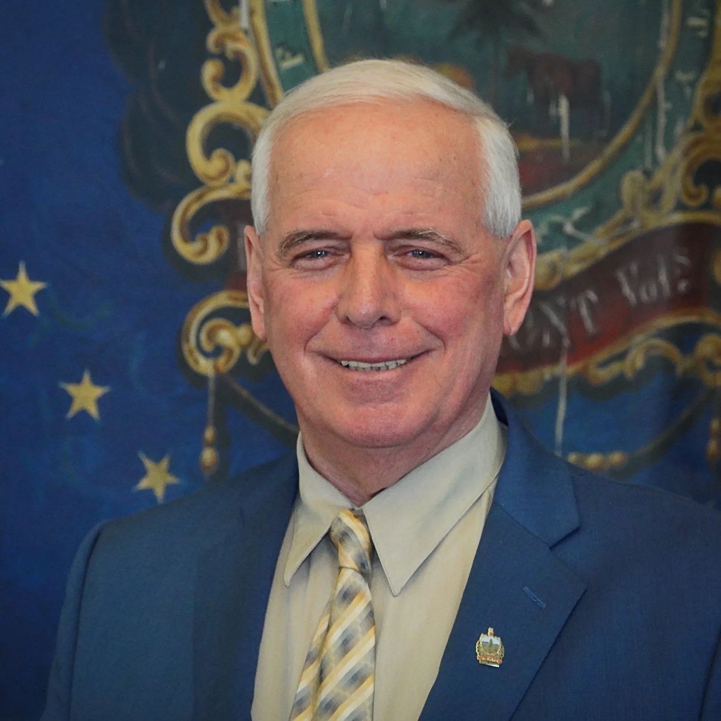 A smiling man with white hair wearing a blue suit, a light-colored shirt, and a yellow-striped tie, standing in front of a background with a coat of arms and stars.