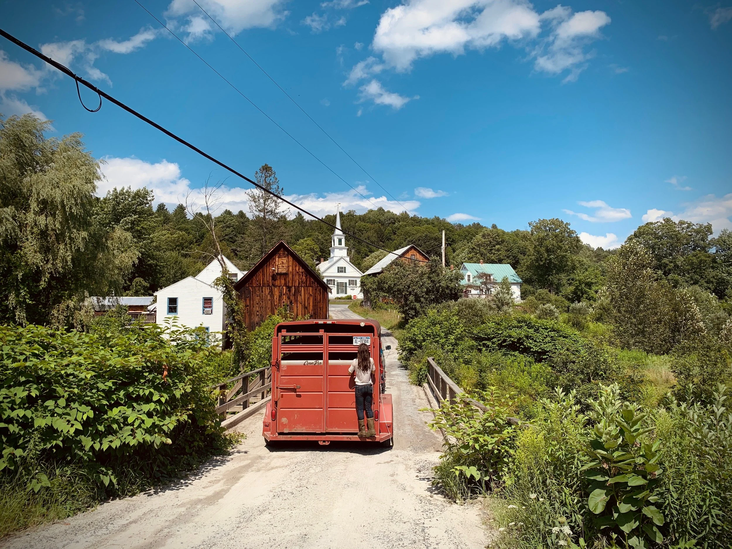 A person riding in the back of a red truck on a dirt road through a rural area with trees and houses, the sky is blue with some clouds.