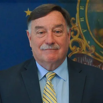 A middle-aged man with gray hair and a mustache, wearing a dark suit, light blue shirt, and striped yellow tie, standing in front of a blue and gold background with an emblem.
