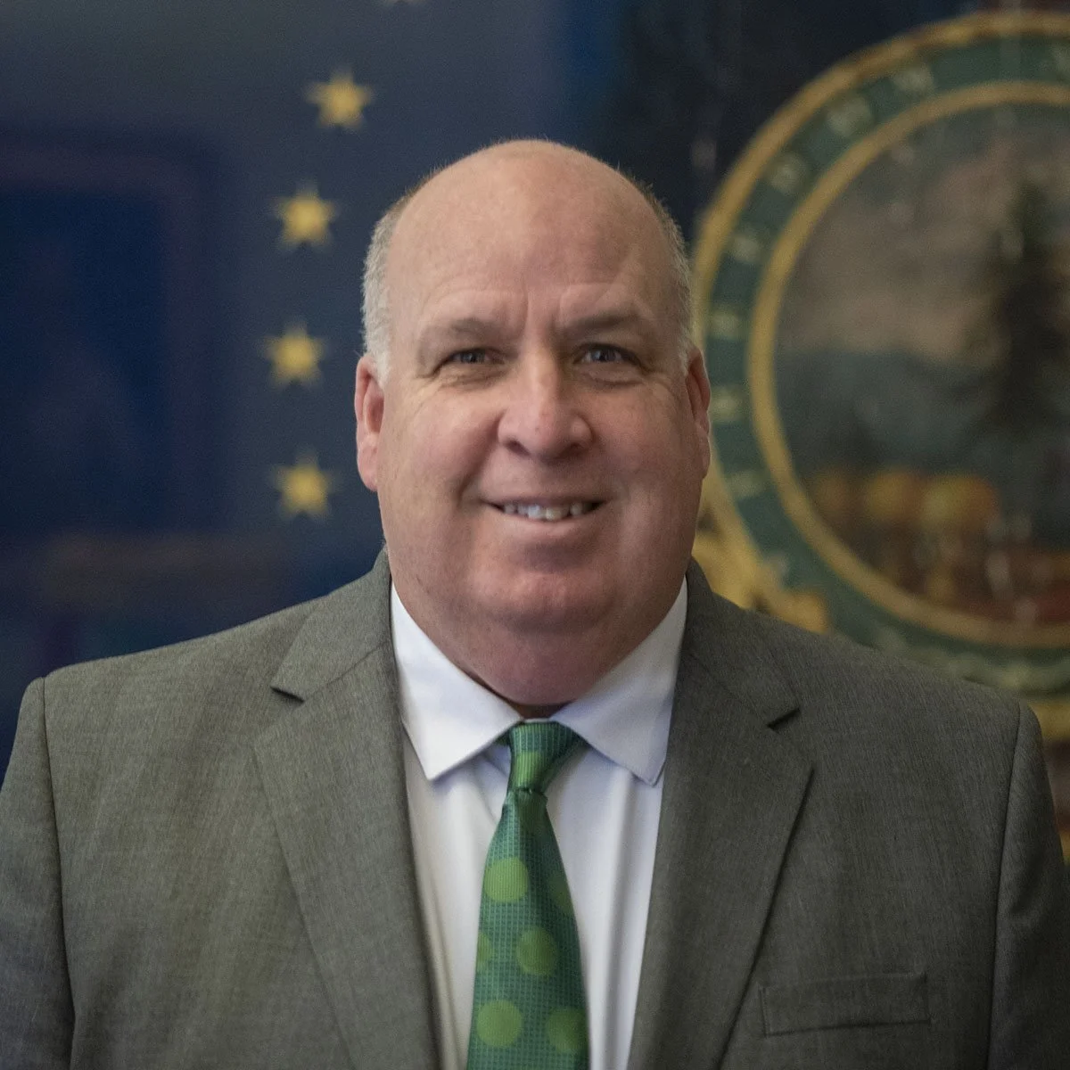 A man wearing a gray suit, white shirt, and green tie, smiling in front of a background with the U.S. flag and a circular emblem.