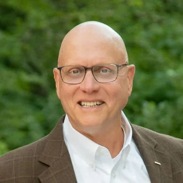 A bald man with glasses wearing a white shirt and brown blazer, smiling outdoors with green trees in the background.