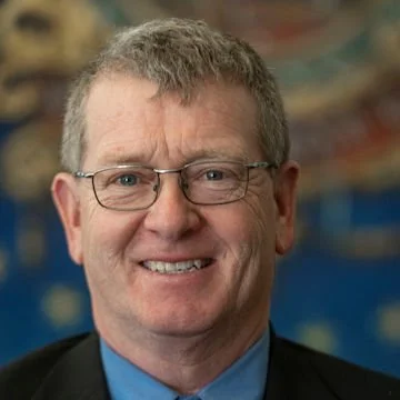 A middle-aged man with glasses, short gray hair, and a friendly smile, wearing a dark suit and blue shirt, standing in front of a colorful background.