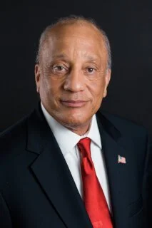 Portrait of an older African American man in a dark suit, white shirt, red tie, and American flag pin on lapel.