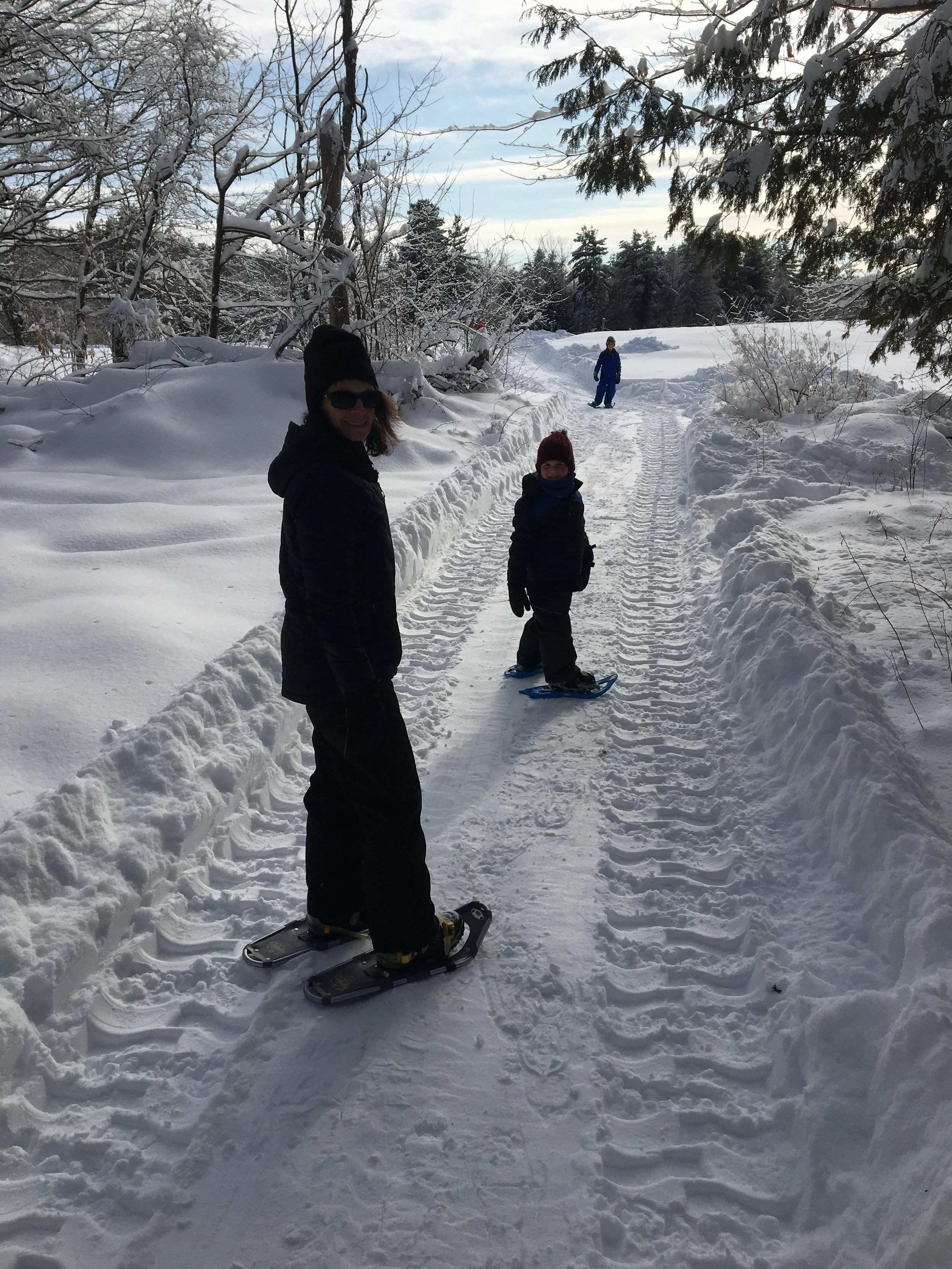 A family enjoying a snowy landscape outdoors during daytime. Two children and a woman are visible; one child is on a snowboard, and another child and woman are smiling at the camera. The snow-covered ground is flanked by trees and bushes, with some branches laden with snow. The sky is partly cloudy.