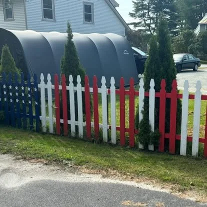 A row of American flag-themed and red and white picket fences, with small green trees behind them, in a residential yard.