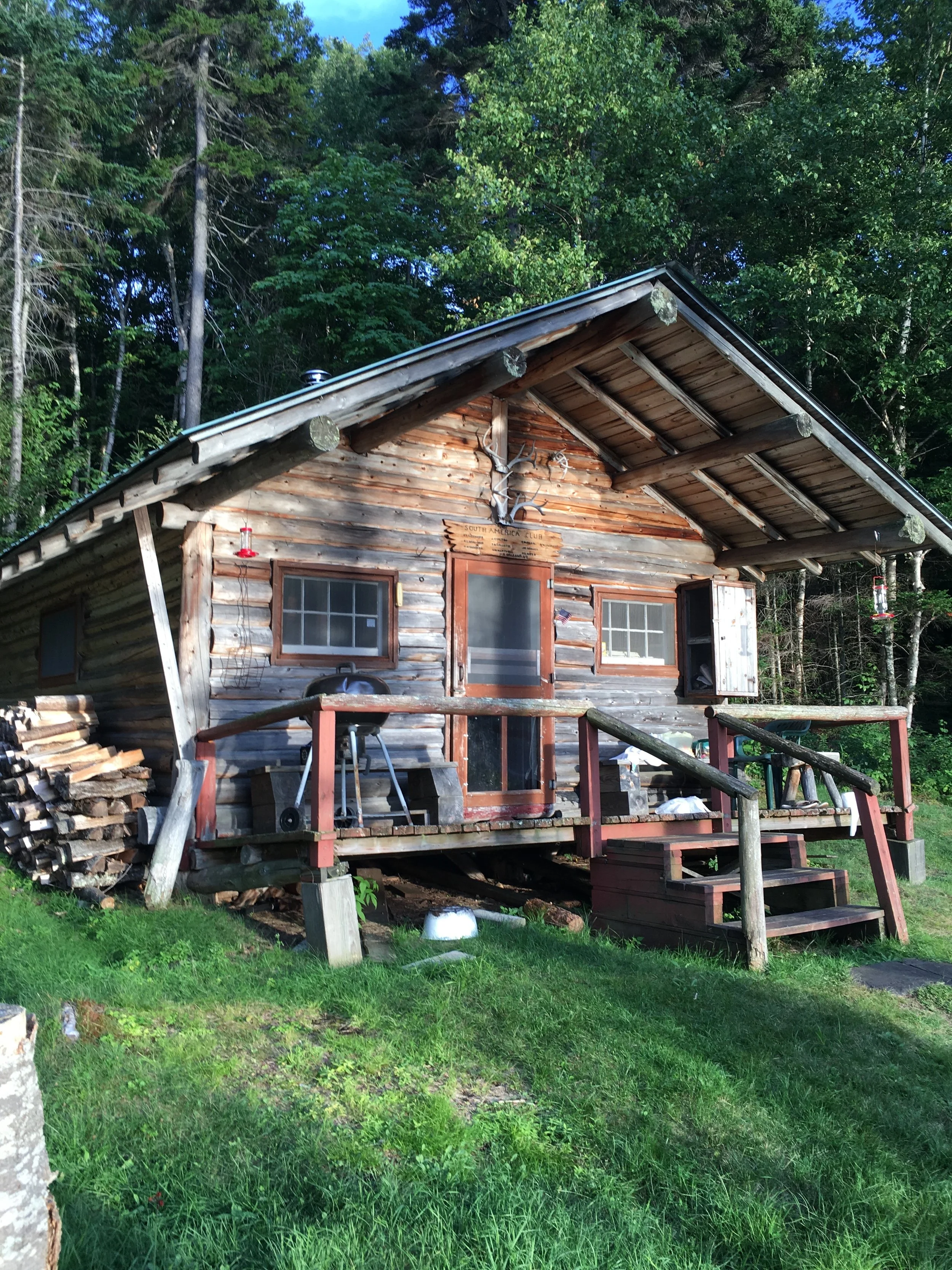 A rustic wooden cabin with a sloped metal roof, set in a forested area. The cabin has two small windows and a door, with steps leading up to a small porch. There is a wooden railing around the porch and some firewood piled on the side. The background features tall trees and lush green foliage.