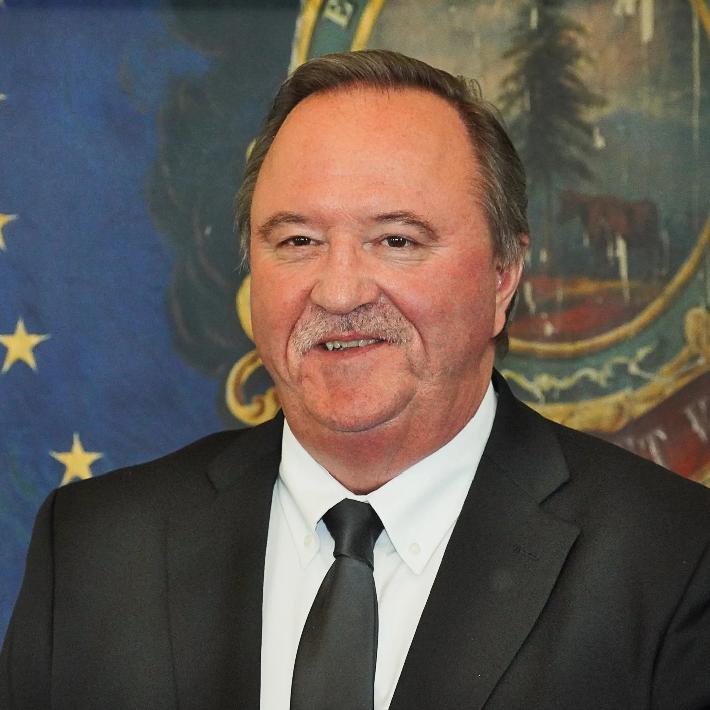 A man with dark hair and a mustache wearing a black suit, white shirt, and black tie, standing in front of a background with the American flag and a state seal.