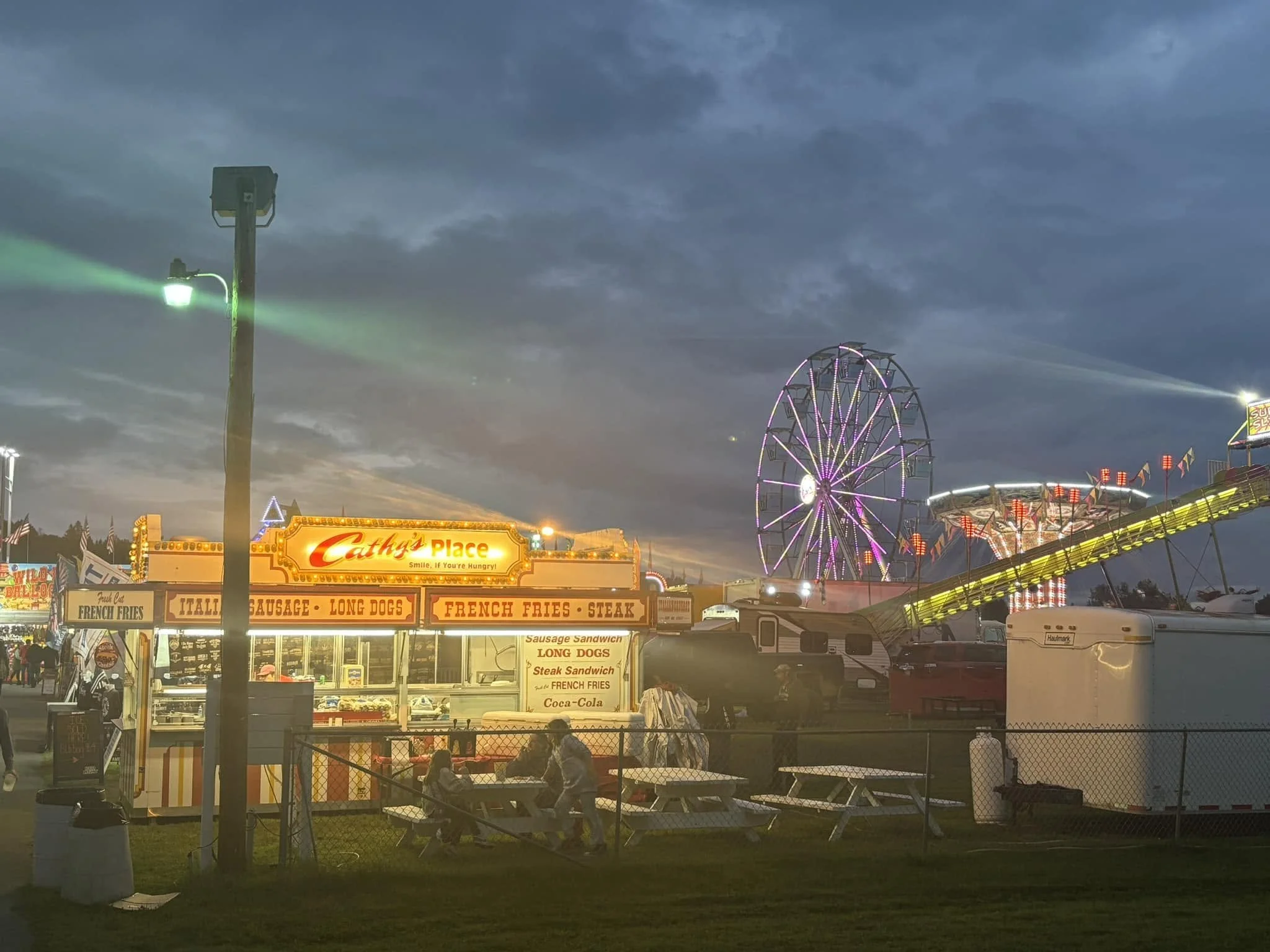 A carnival scene at dusk with a large Ferris wheel lit up in purple, a roller coaster with bright yellow and purple lights, and a food stand named Cathy’s Place selling fries, sausage, long dogs, and soda. The sky is cloudy and darkening, and people are sitting and walking around.