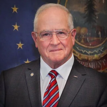 Smiling elderly man in a suit and tie standing in front of a flag with stars.