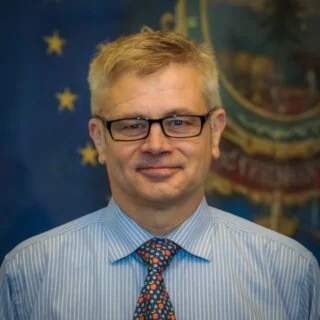 A man with blonde hair, glasses, wearing a blue striped shirt and a colorful tie, standing in front of a decorated blue background with stars and a crest.