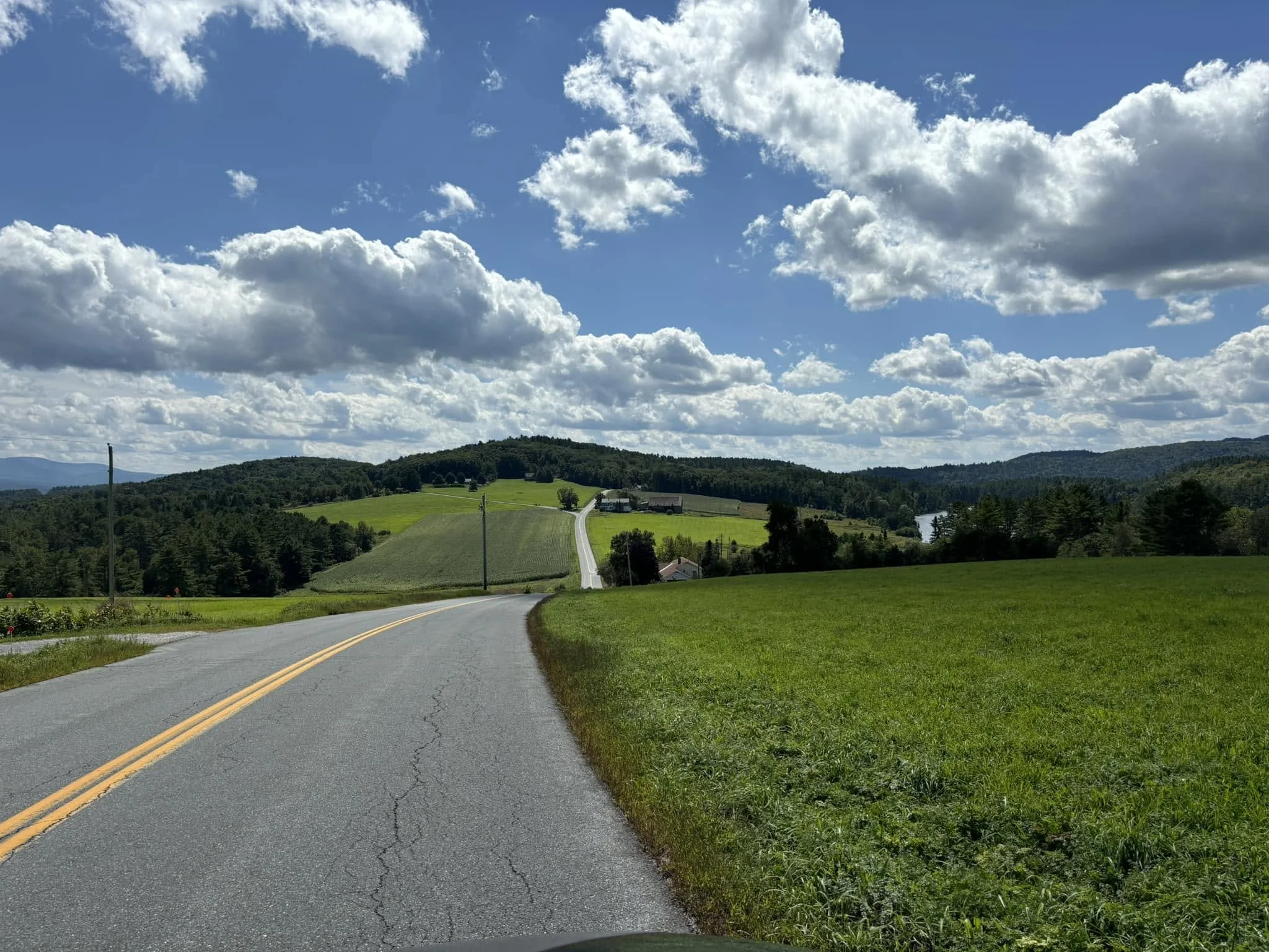 A rural road with a double yellow line leading into green farmland, trees, and hills under a partly cloudy sky.