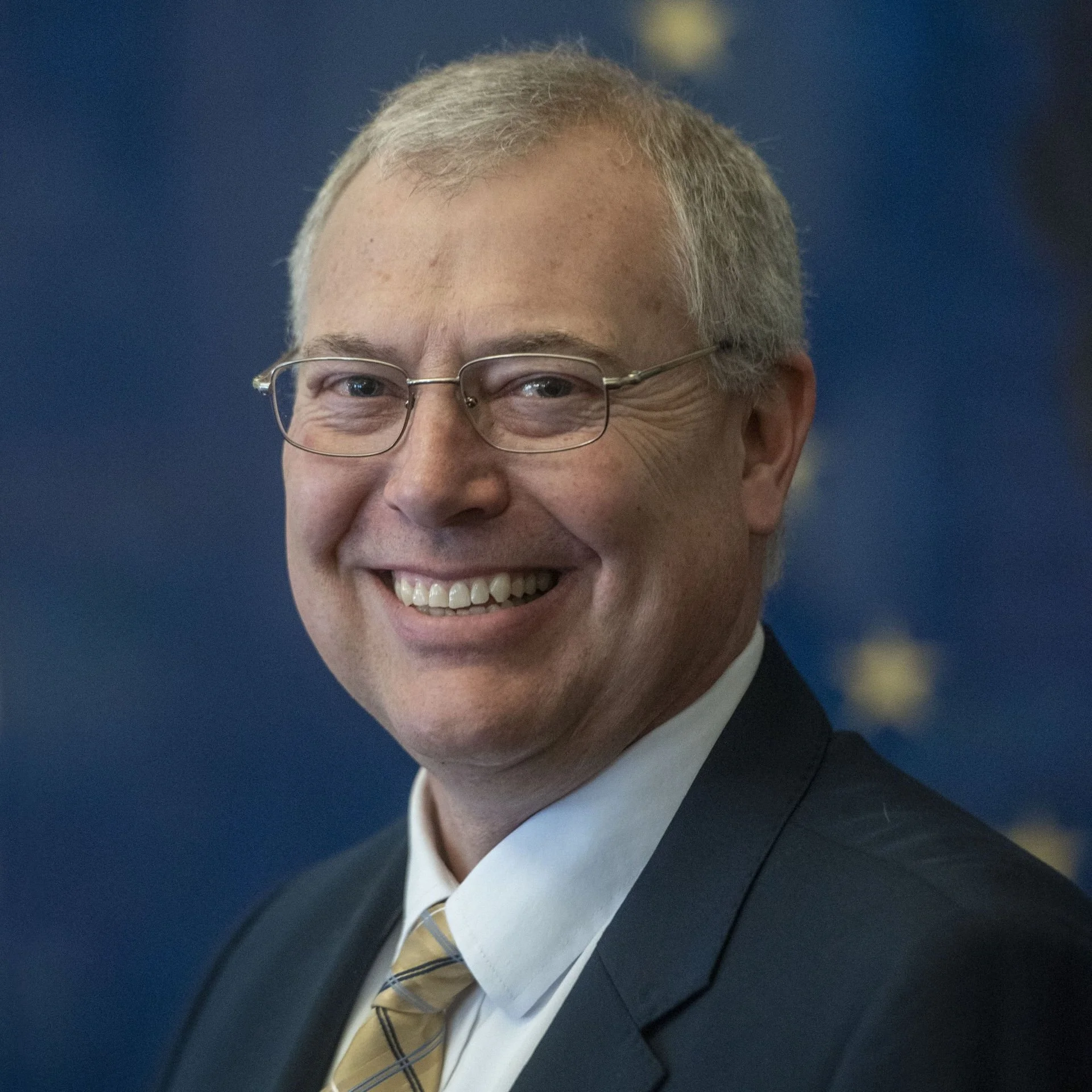 Close-up of a smiling man wearing glasses, a navy blazer, a white shirt, and a yellow plaid tie, with a blurred dark blue background.