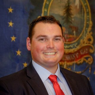A young man with short dark hair, wearing a suit with a light blue shirt and a red tie, smiling in front of a background featuring a U.S. state emblem and a starry blue backdrop.