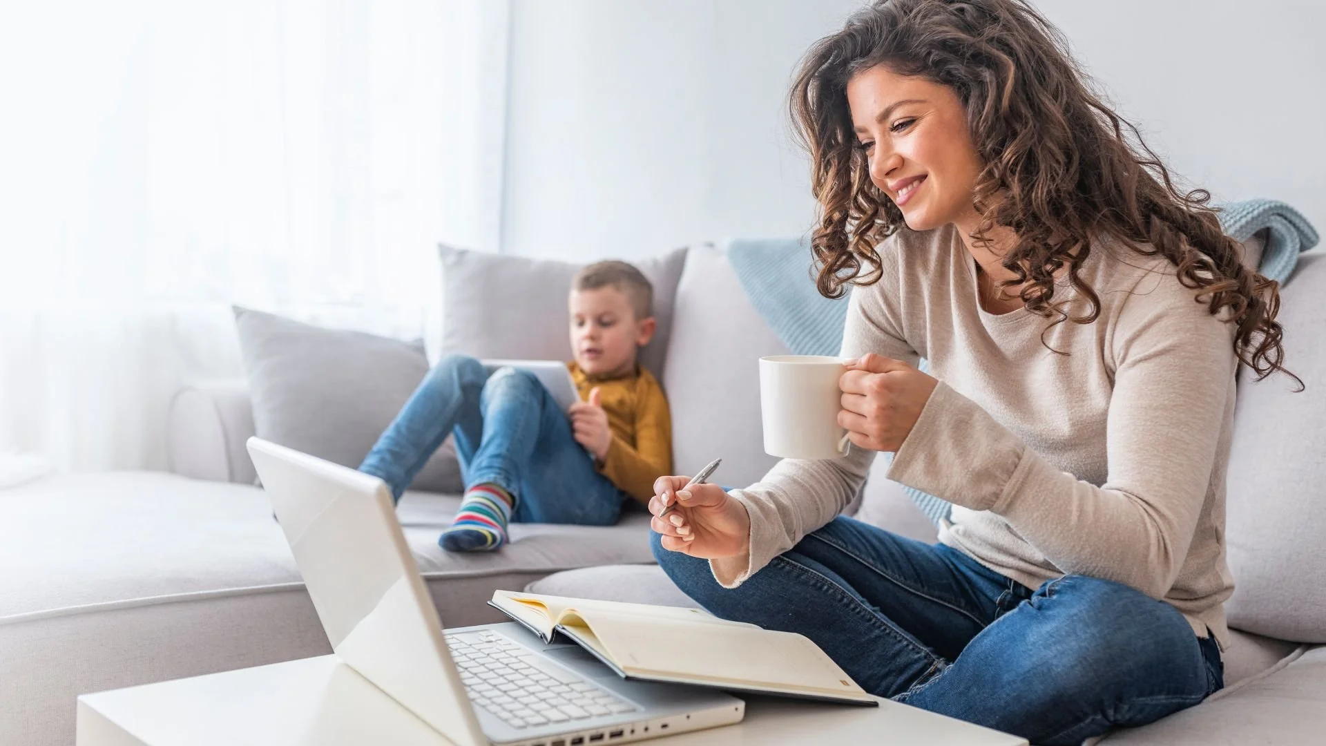 A woman with curly hair sitting on a couch, holding a white mug, smiling, with a child in a yellow shirt and blue jeans sitting behind her on the couch, using a tablet.