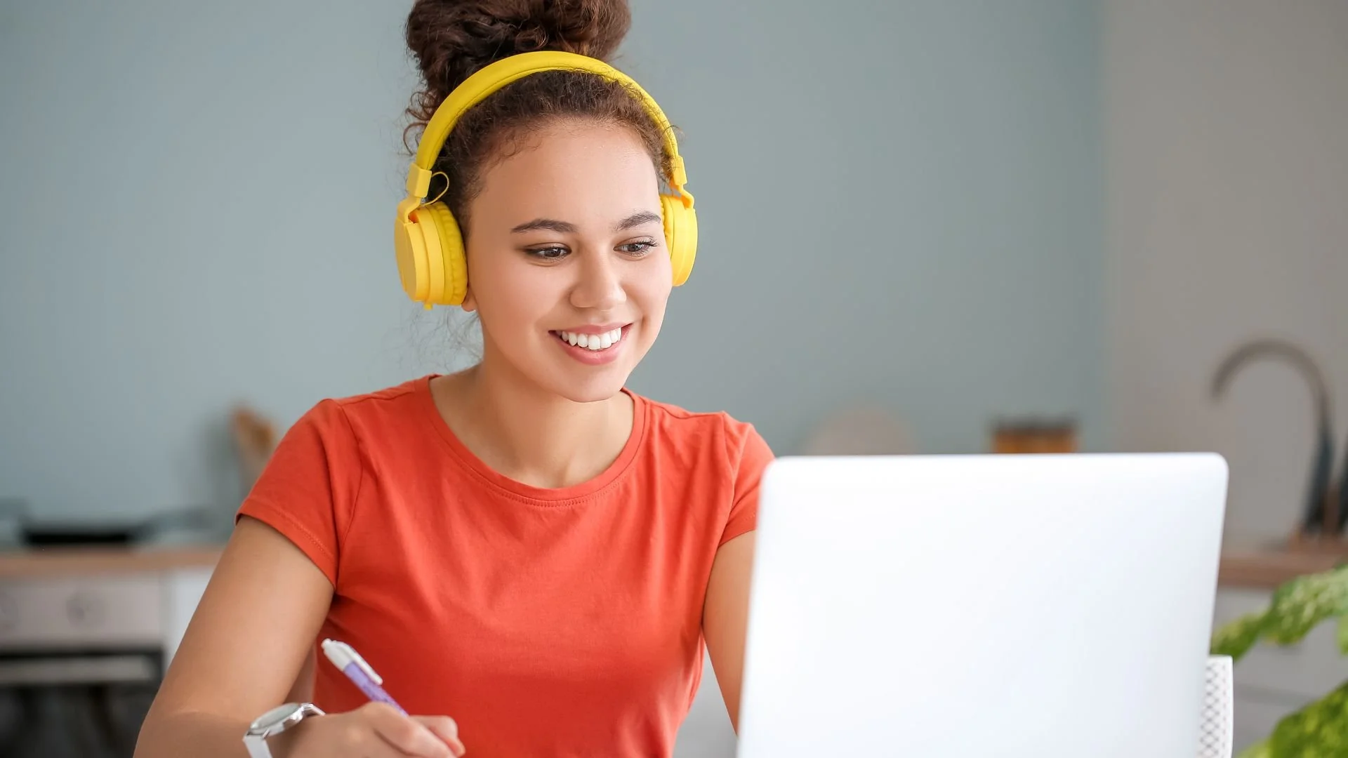 A young woman with curly hair in a top bun, wearing yellow headphones and an orange t-shirt, working on a laptop in a kitchen or home office setting.