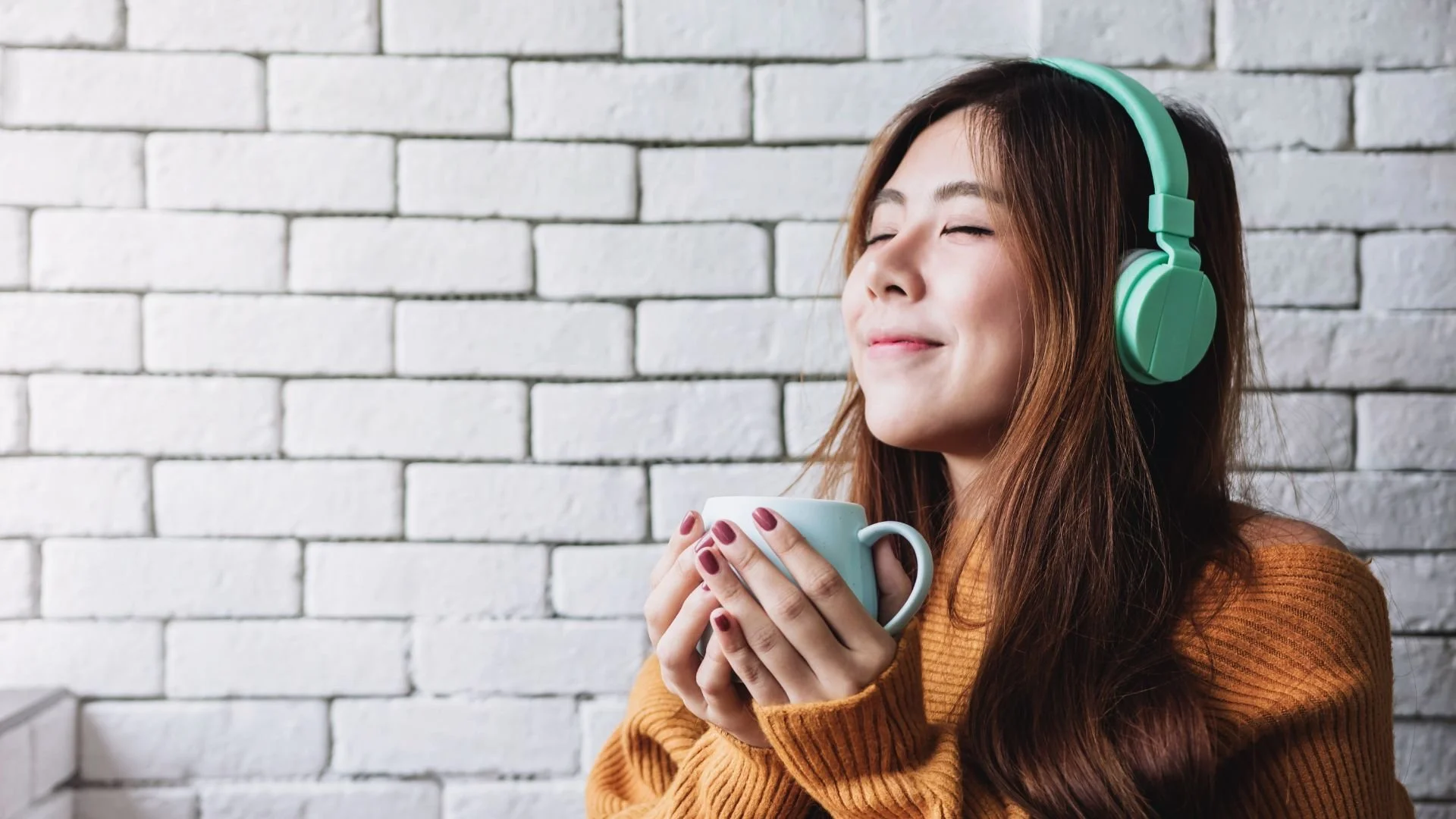 A woman with long brown hair wearing a green headphone is holding a white mug with both hands, smiling with her eyes closed, standing in front of a white brick wall.
