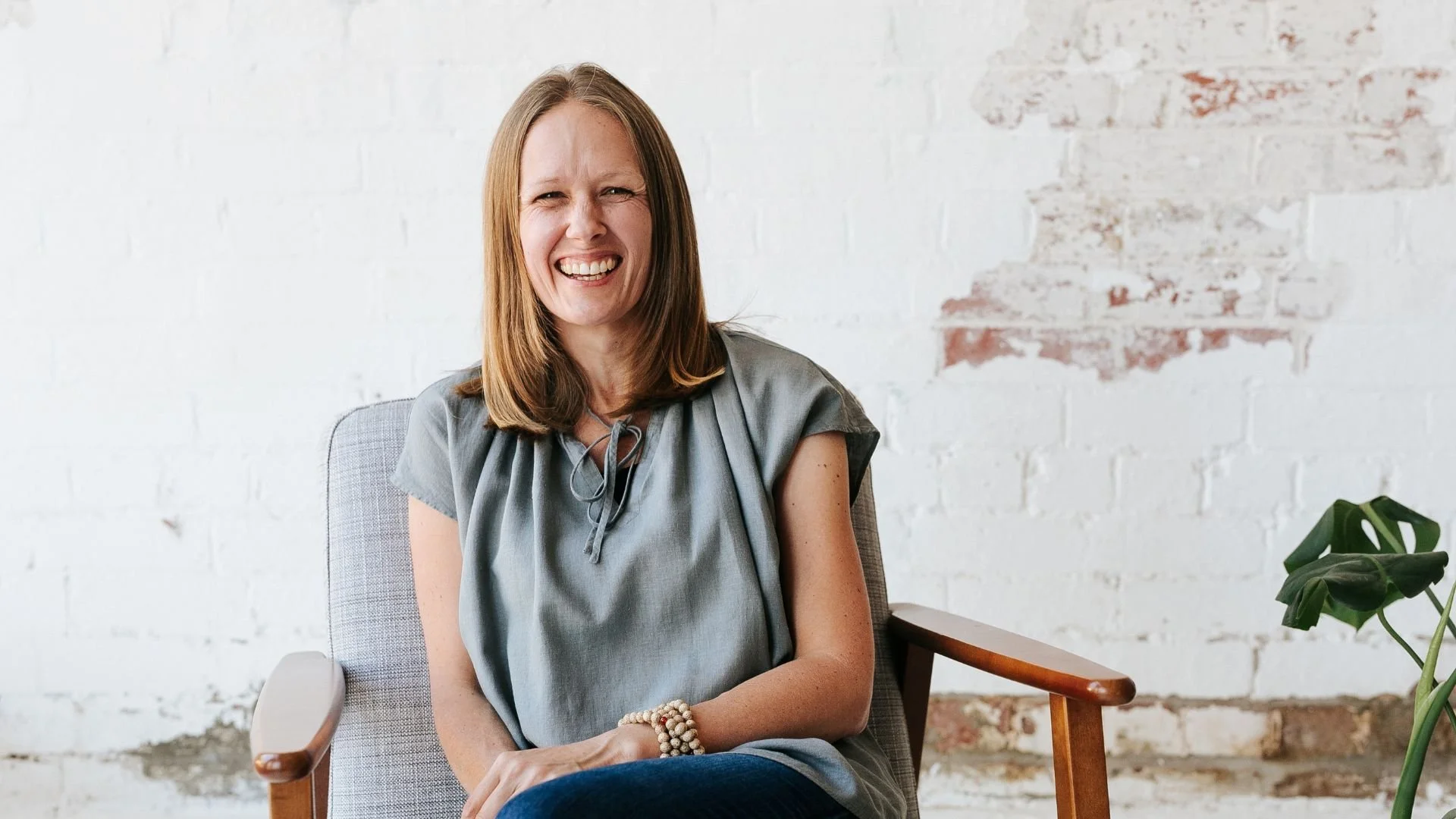 A woman with shoulder-length red hair smiling and sitting in a modern armchair against a white brick wall with some peeling paint, wearing a gray short-sleeved top and a beaded bracelet.