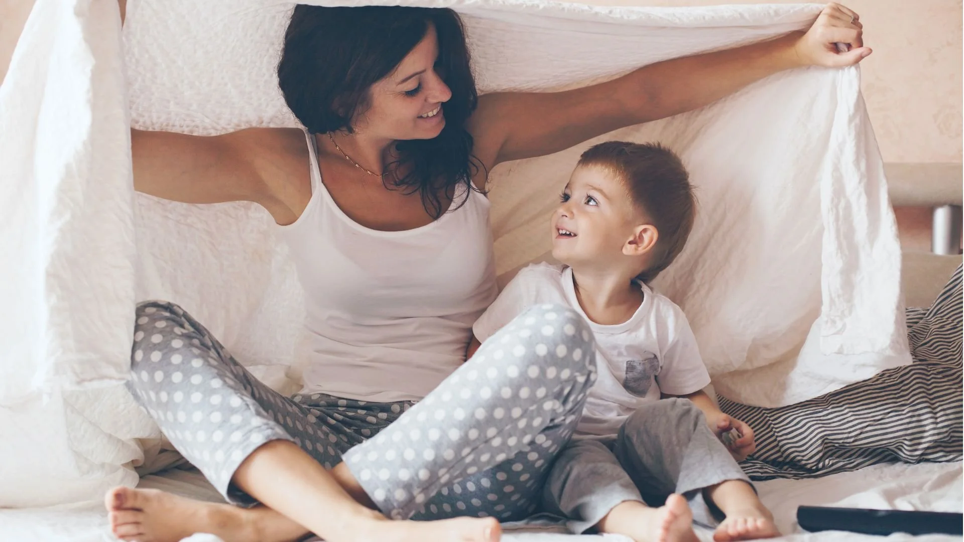 A woman and a young boy sitting inside a makeshift tent made of bedsheets, smiling and looking at each other.