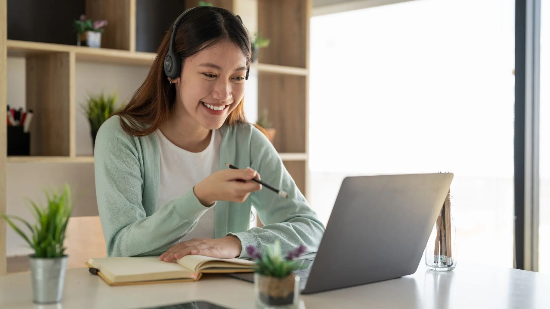 Young woman wearing headphones, smiling, at a workspace with a laptop, open notebook, and potted plants