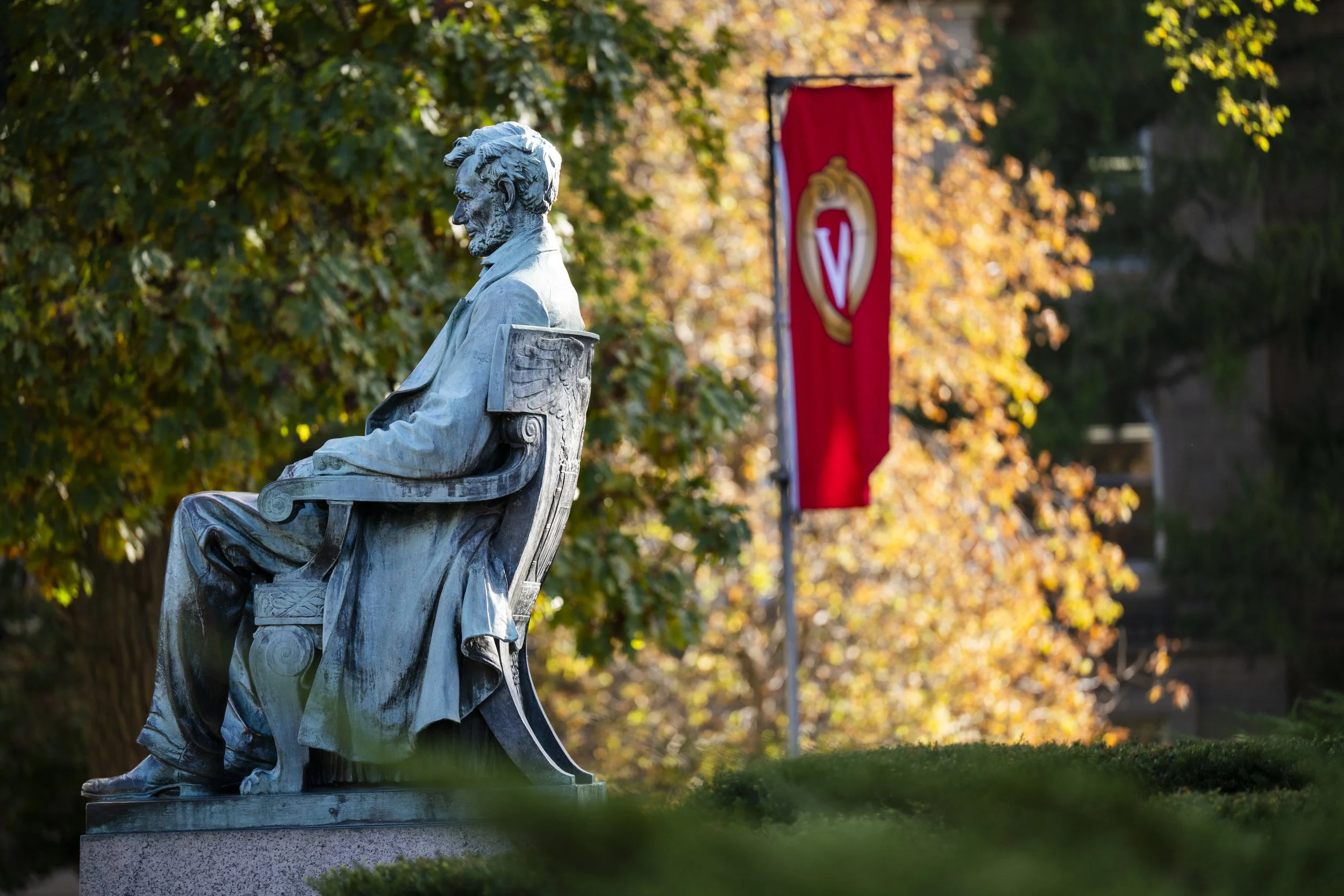Bronze statue of a seated man with a beard, wearing a coat, outdoors in autumn with colorful trees and a red banner in the background.