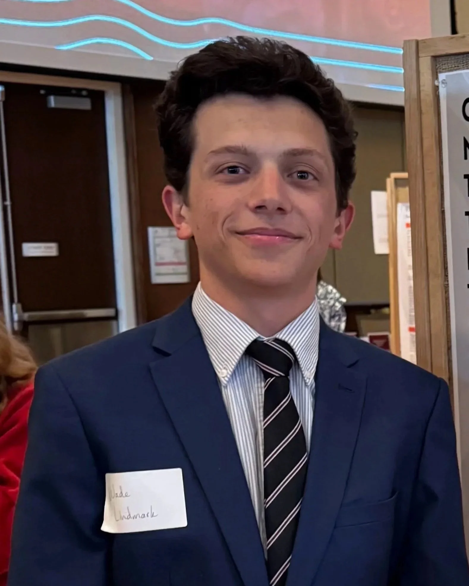 A young man with dark curly hair, wearing a dark blue suit, white shirt, and striped tie, smiling at the camera. He has a paper name tag attached to his suit with handwritten text.