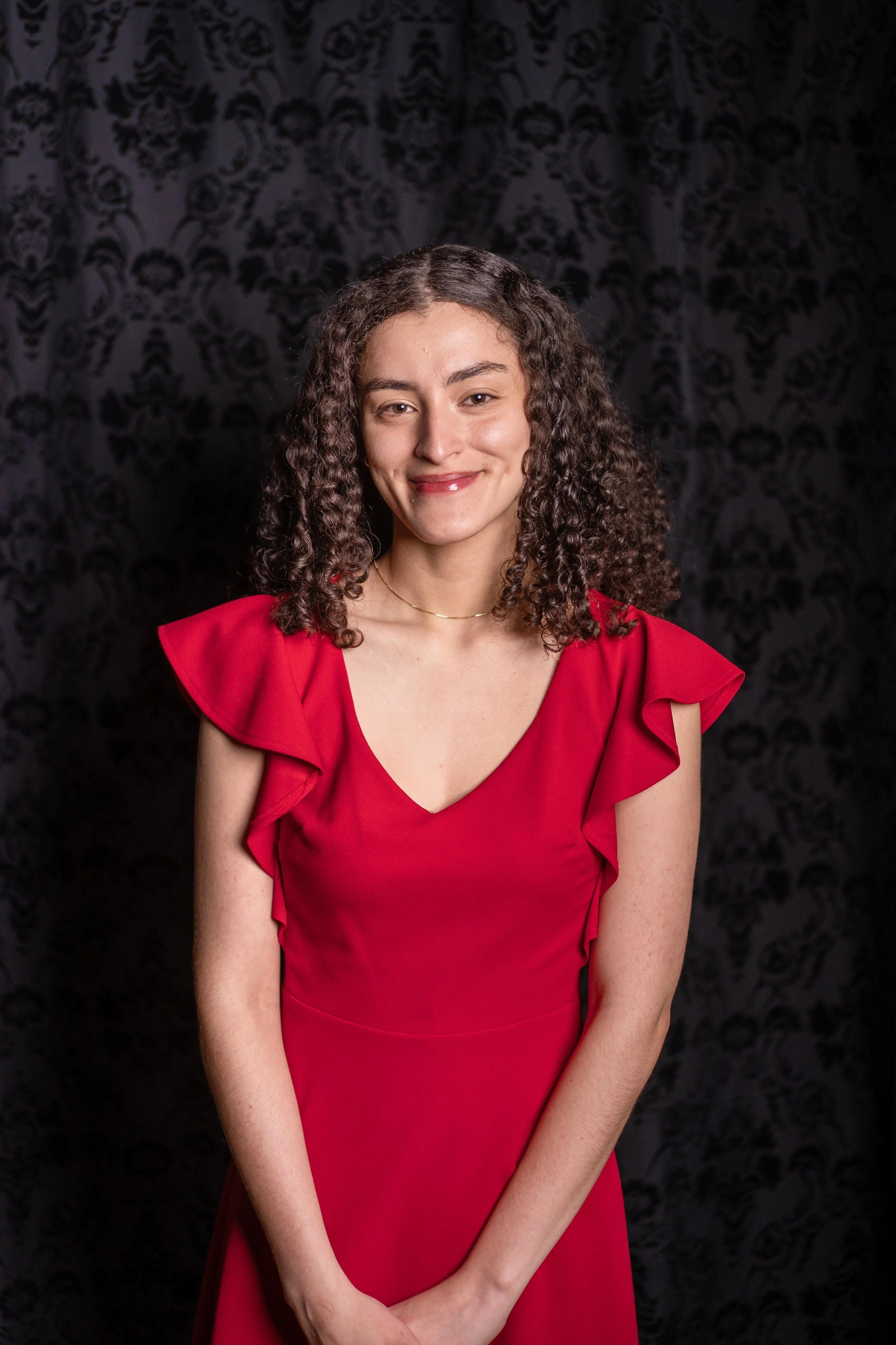A woman with curly hair wearing a red dress, standing against a black patterned background.