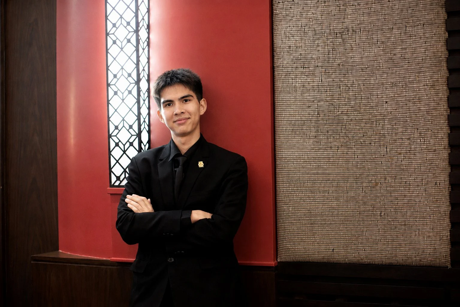 Young man in black suit with arms crossed standing in front of red wall with textured pattern and decorative window.