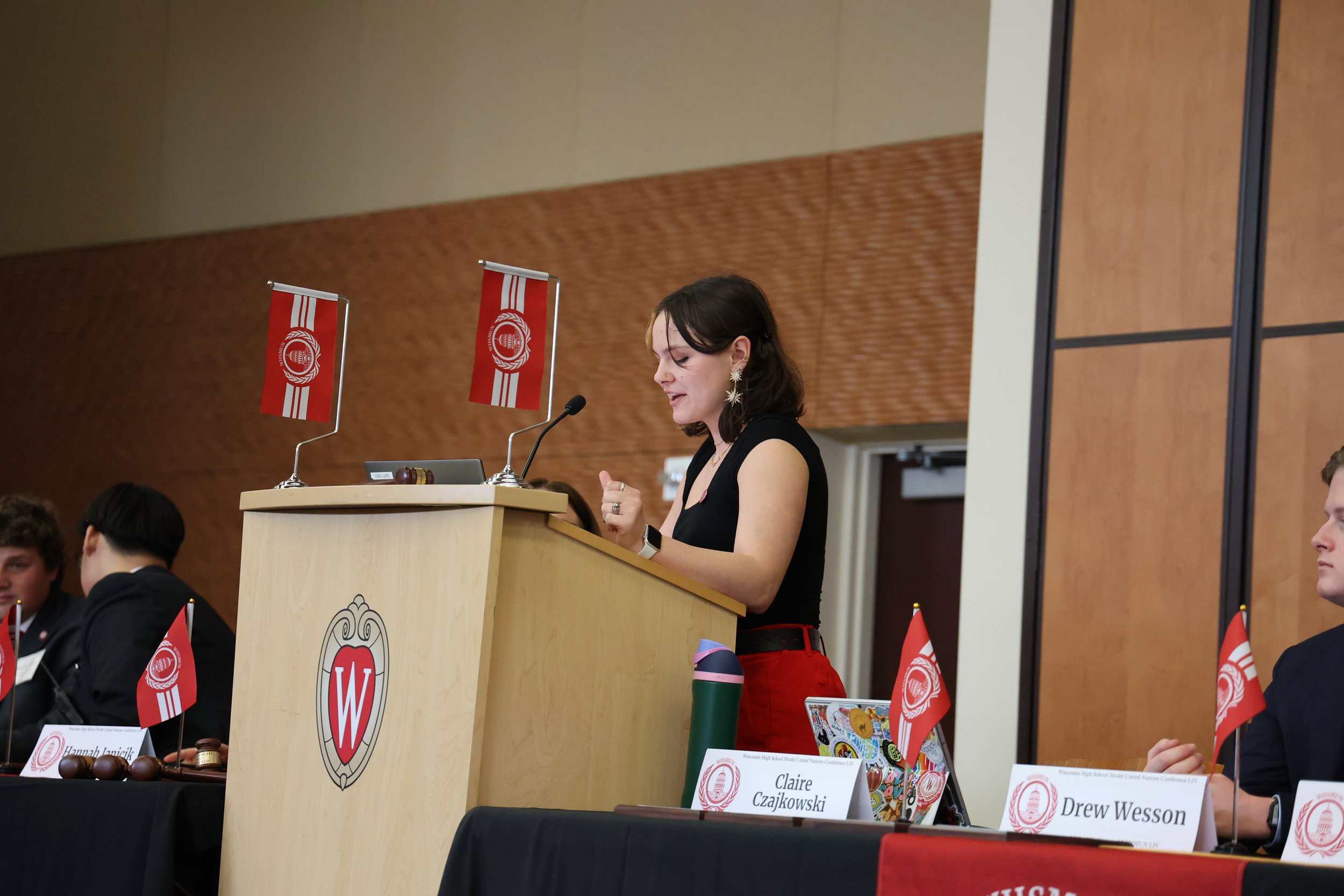 A woman with dark hair and star-shaped earrings stands at a podium, speaking into a microphone during a formal event. Behind her, flags and other individuals are seated at tables with name tags.